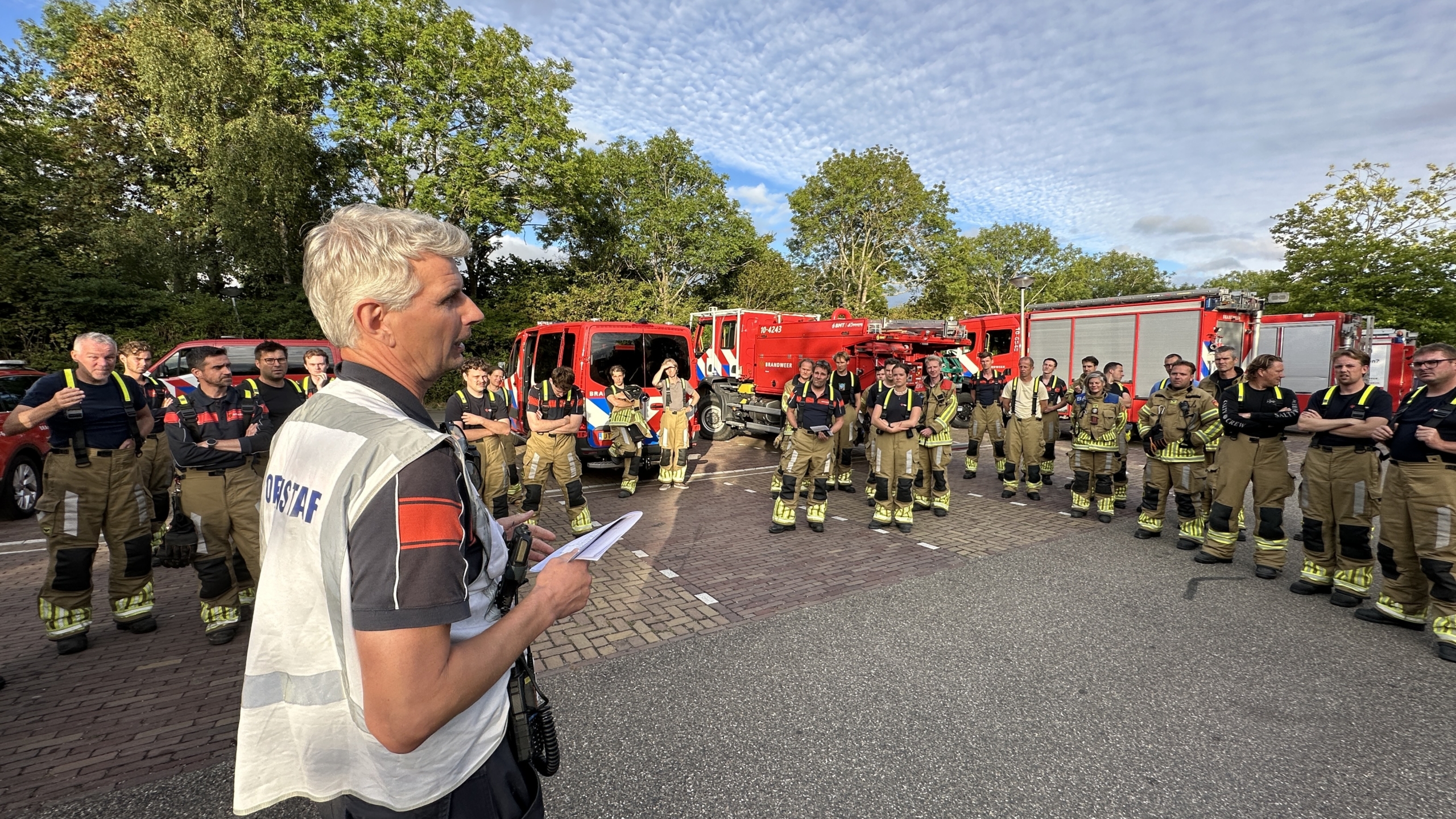 Leider geeft briefing aan groep brandweerlieden, met bluswagens op de achtergrond op een parkeerterrein