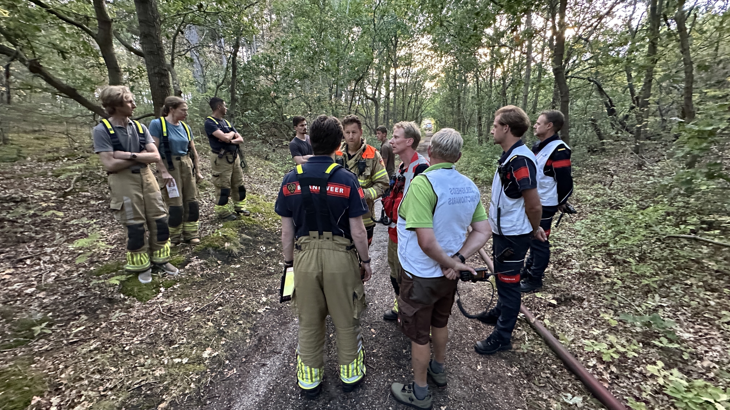 Brandweerlieden en hulpverleners overleggen op een bospad tijdens een inzet, met brandslang langs de weg