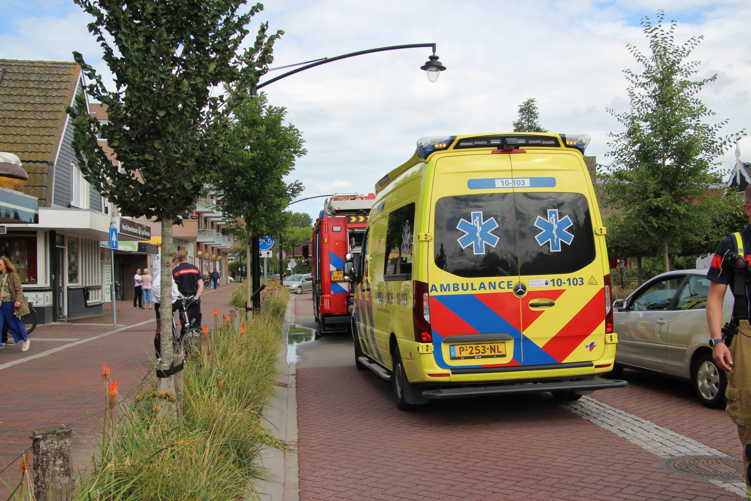 Gele ambulance en rode brandweerwagen geparkeerd op een straat in woonwijk, met hulpverleners en omstanders