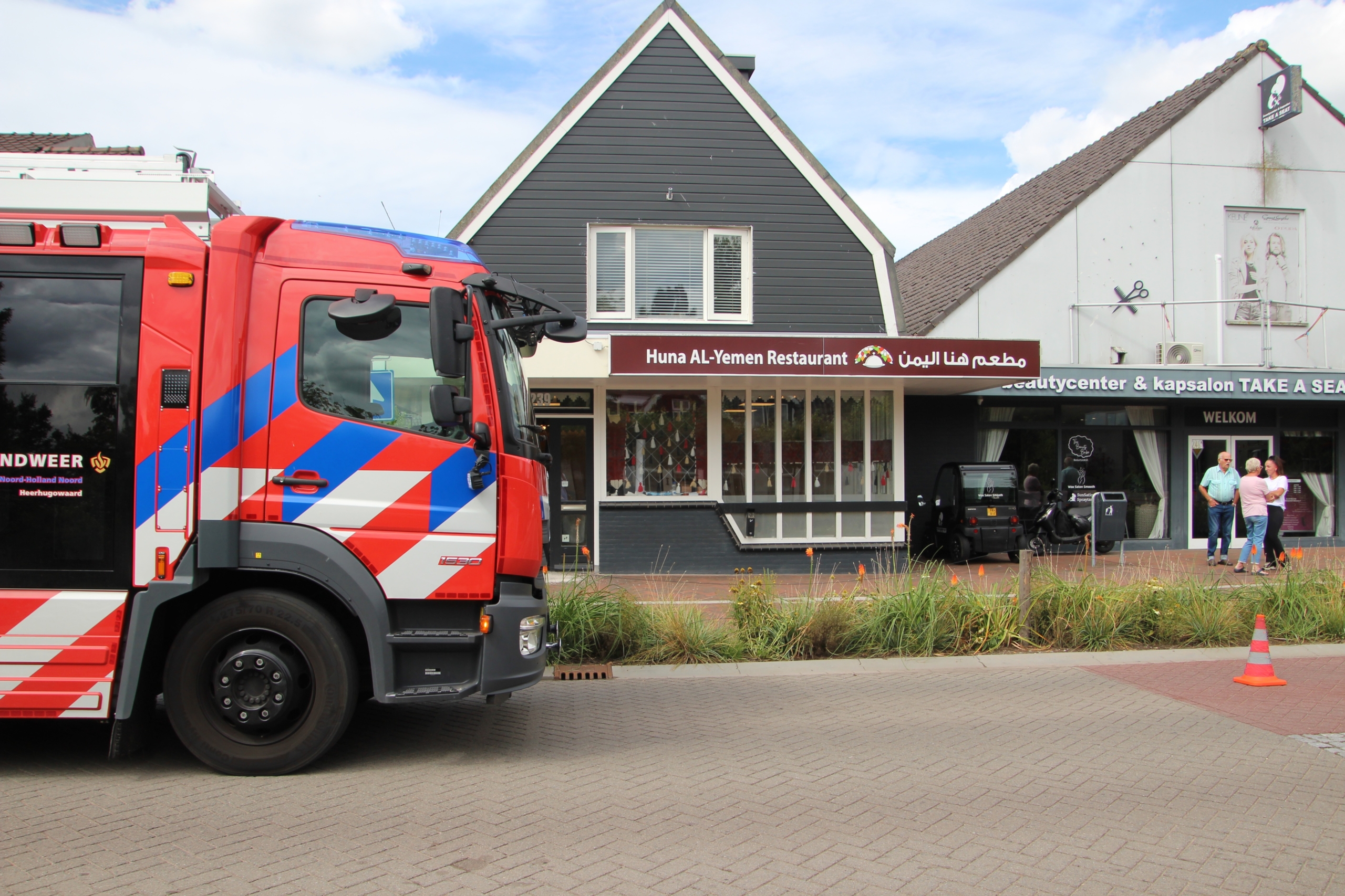 Brandweerwagen geparkeerd voor Huna AL-Yemen Restaurant naast een kapperszaak, met enkele mensen bij de ingang