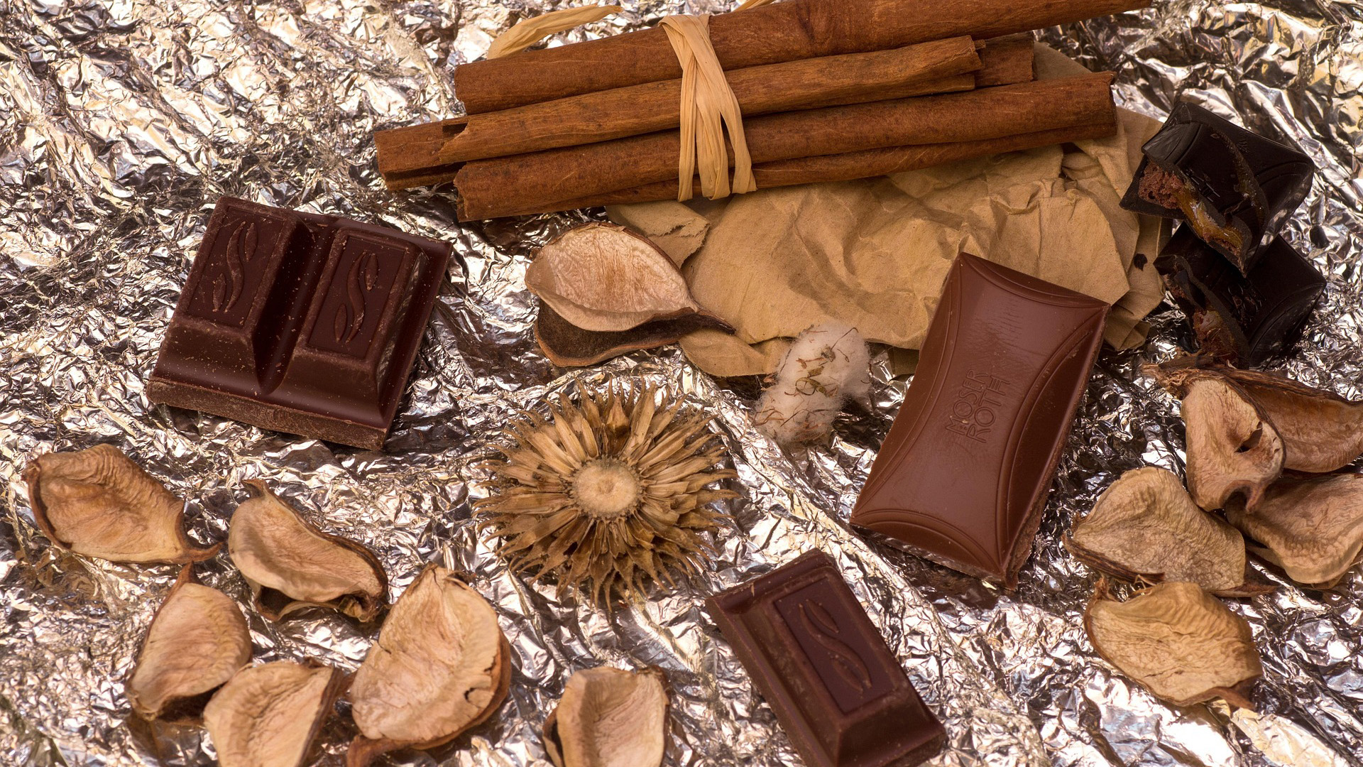 Stukken chocolade en een bundel kaneelstokjes op verfrommeld aluminiumfolie, met gedroogde bloemblaadjes eromheen