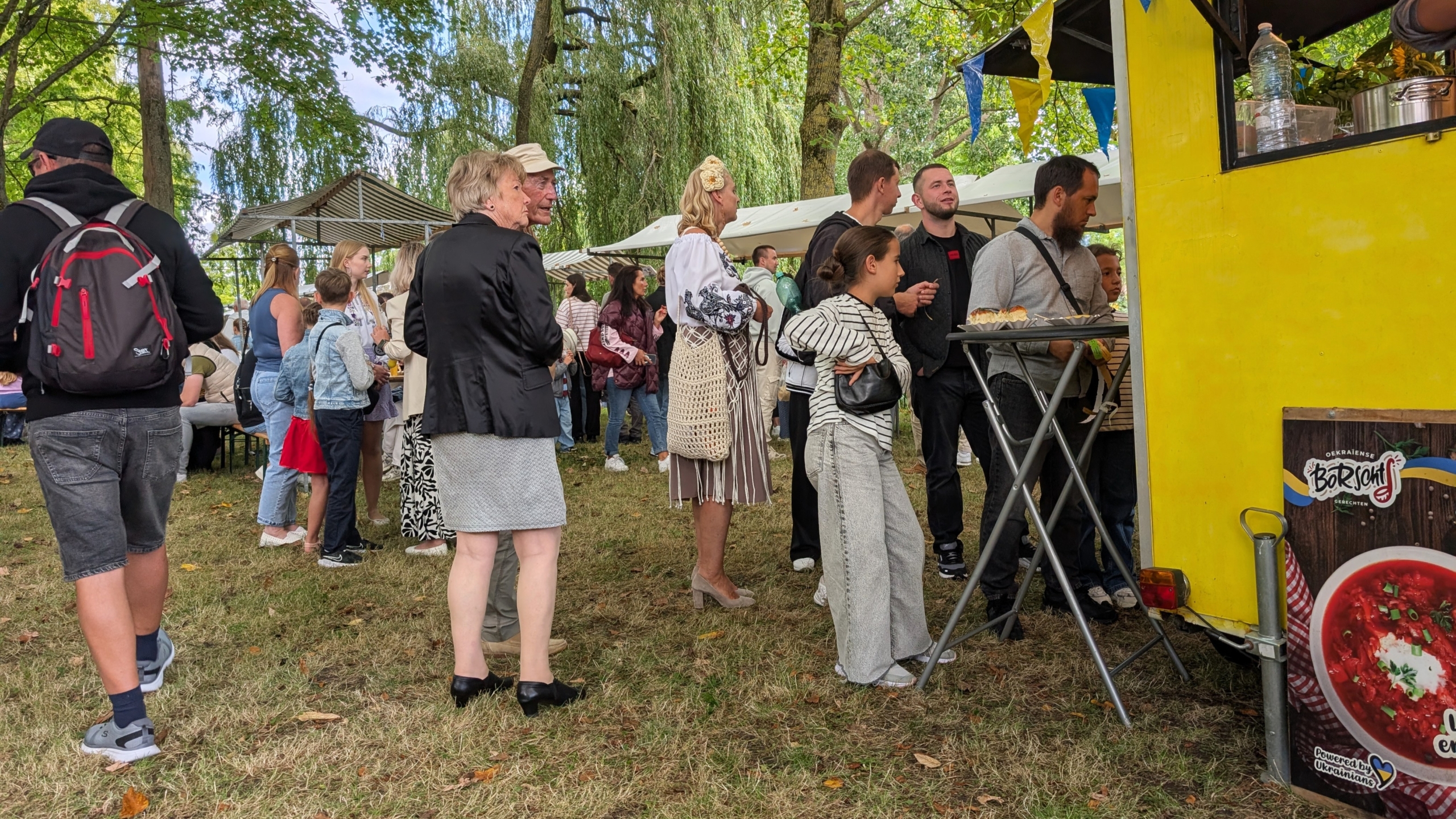 Bezoekers staan in de rij bij een gele foodtruck in een park, met marktkramen en bomen op de achtergrond