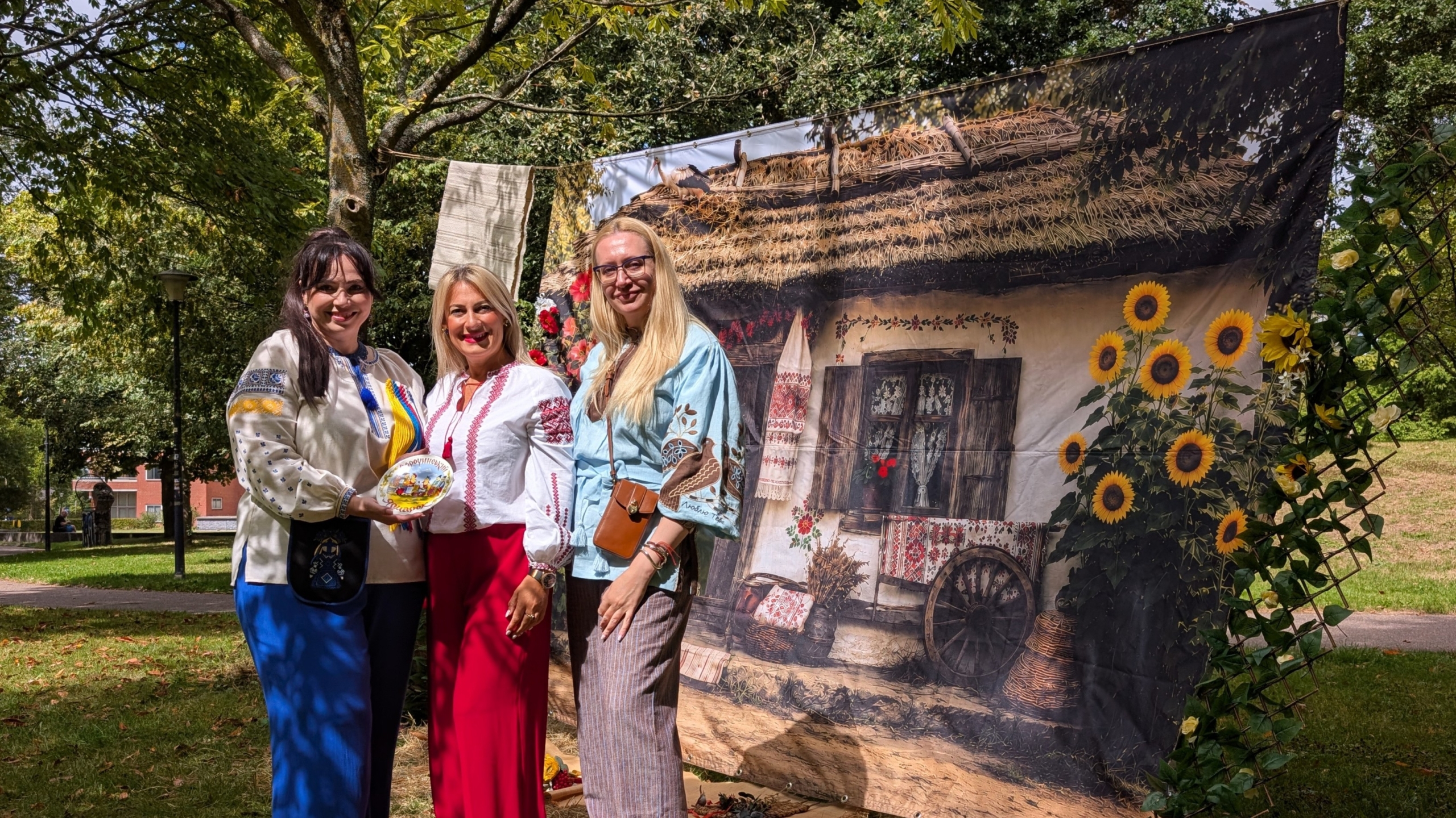 Drie vrouwen in traditionele geborduurde kleding poseren in een park bij een decor van een boerderij met zonnebloemen