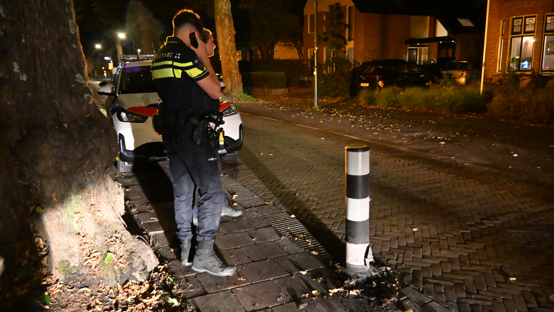 Politieagent telefoneert naast een politieauto op een donkere woonstraat, met een beschadigde paal op de voorgrond