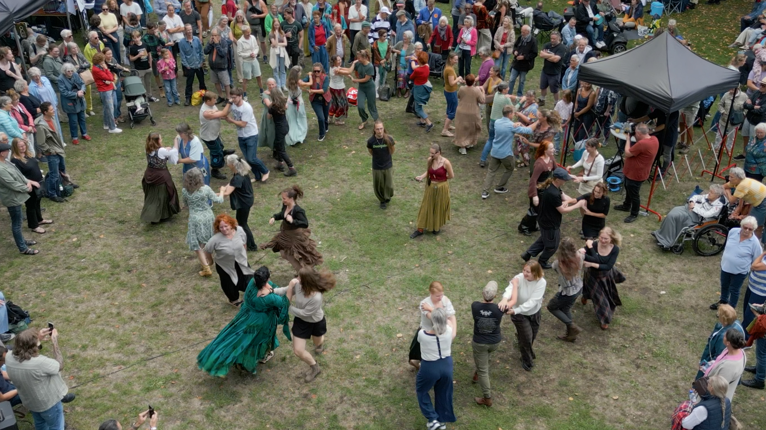 Mensen dansen in paren op een grasveld tijdens een buitenfestival, omringd door toeschouwers en een tent met apparatuur