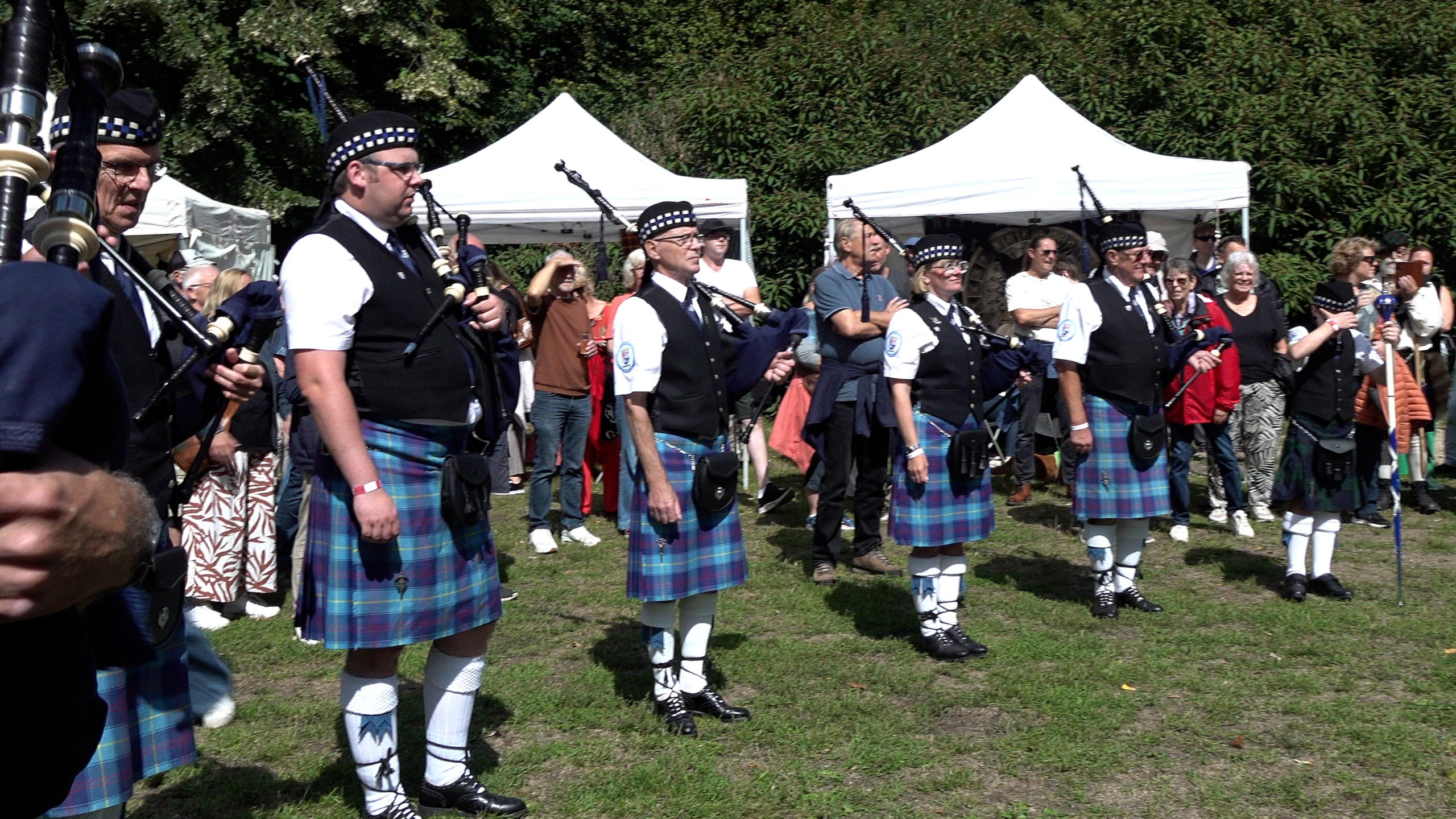 Doedelzakband in kilts speelt buiten op een festival, met publiek en witte tenten op de achtergrond