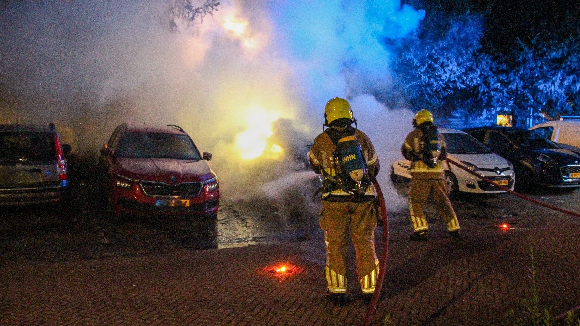 Brandweerlieden blussen brandende auto op parkeerplaats, met veel rook tussen geparkeerde auto’s in het donker