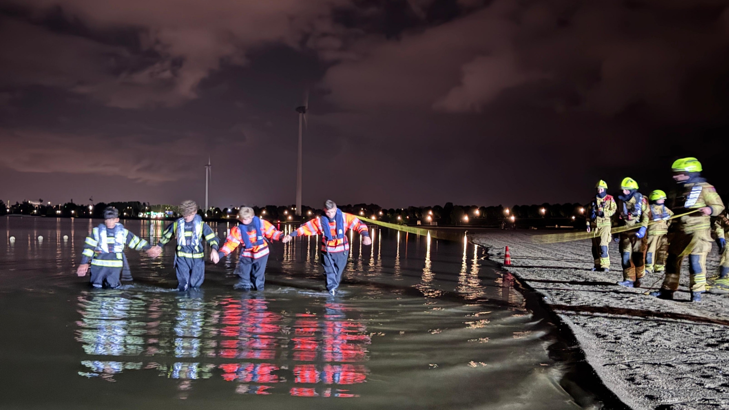 Hulpverleners waden ’s nachts door ondiep water, vast aan een touw, terwijl collega’s op de oever begeleiden