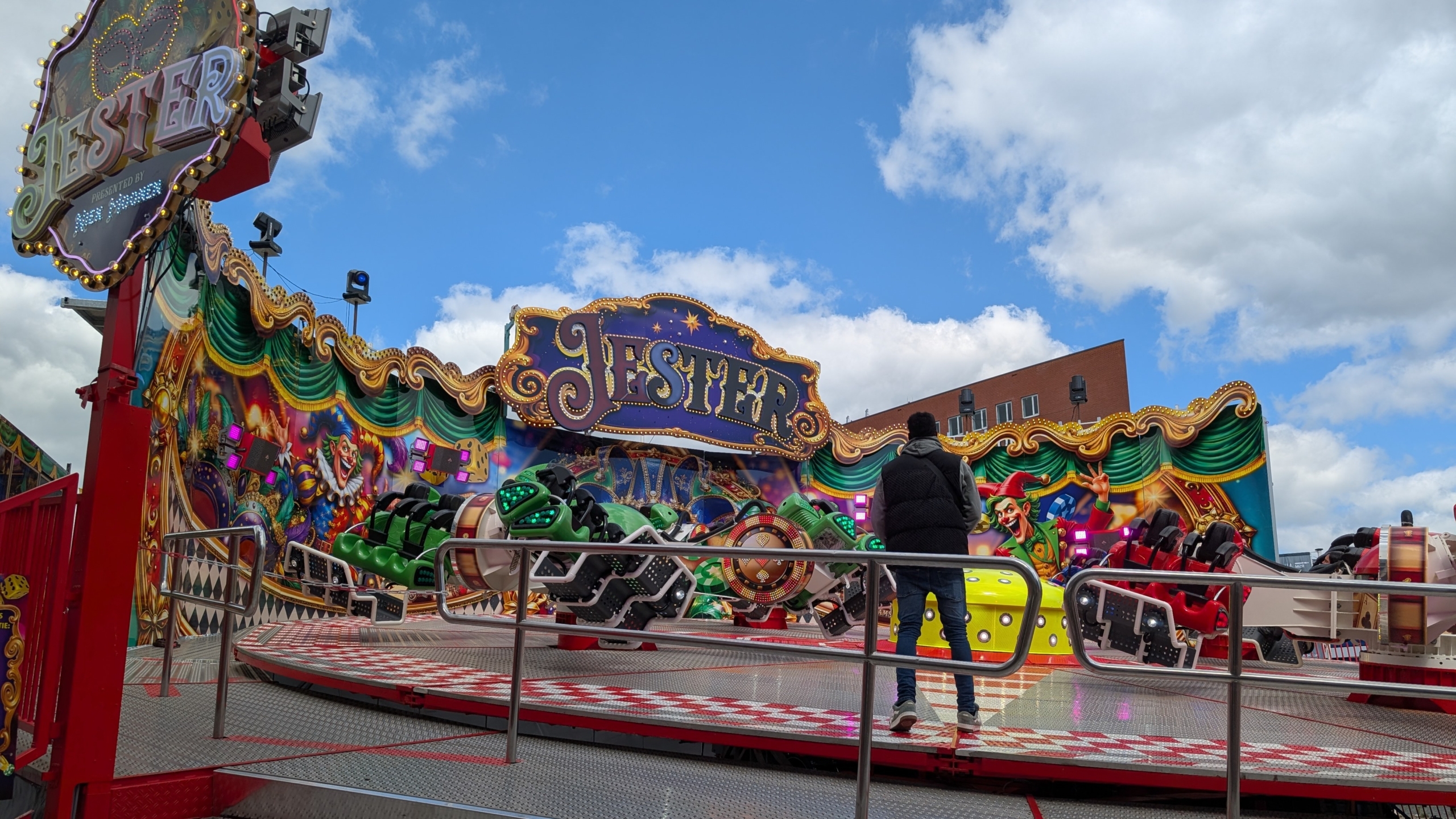 Kermisattractie met bord “Jester” en kleurrijke clownschilderingen, met draaiende gondels en een toeschouwer op het platform