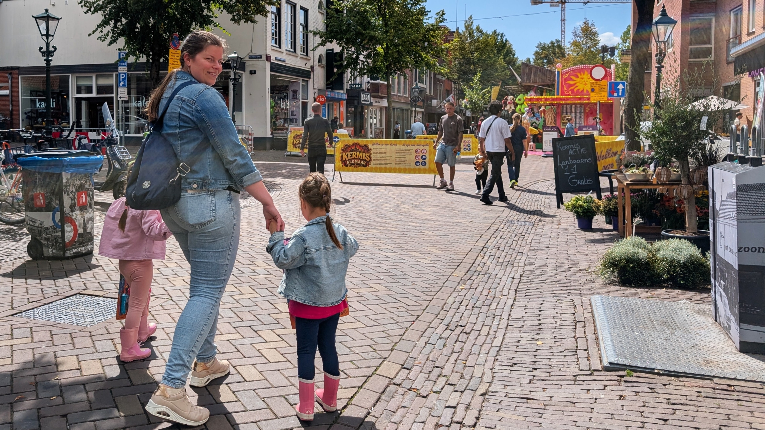 Vrouw met twee kinderen loopt over een geplaveide winkelstraat richting afzetting voor de kermis, met attracties op de achtergrond