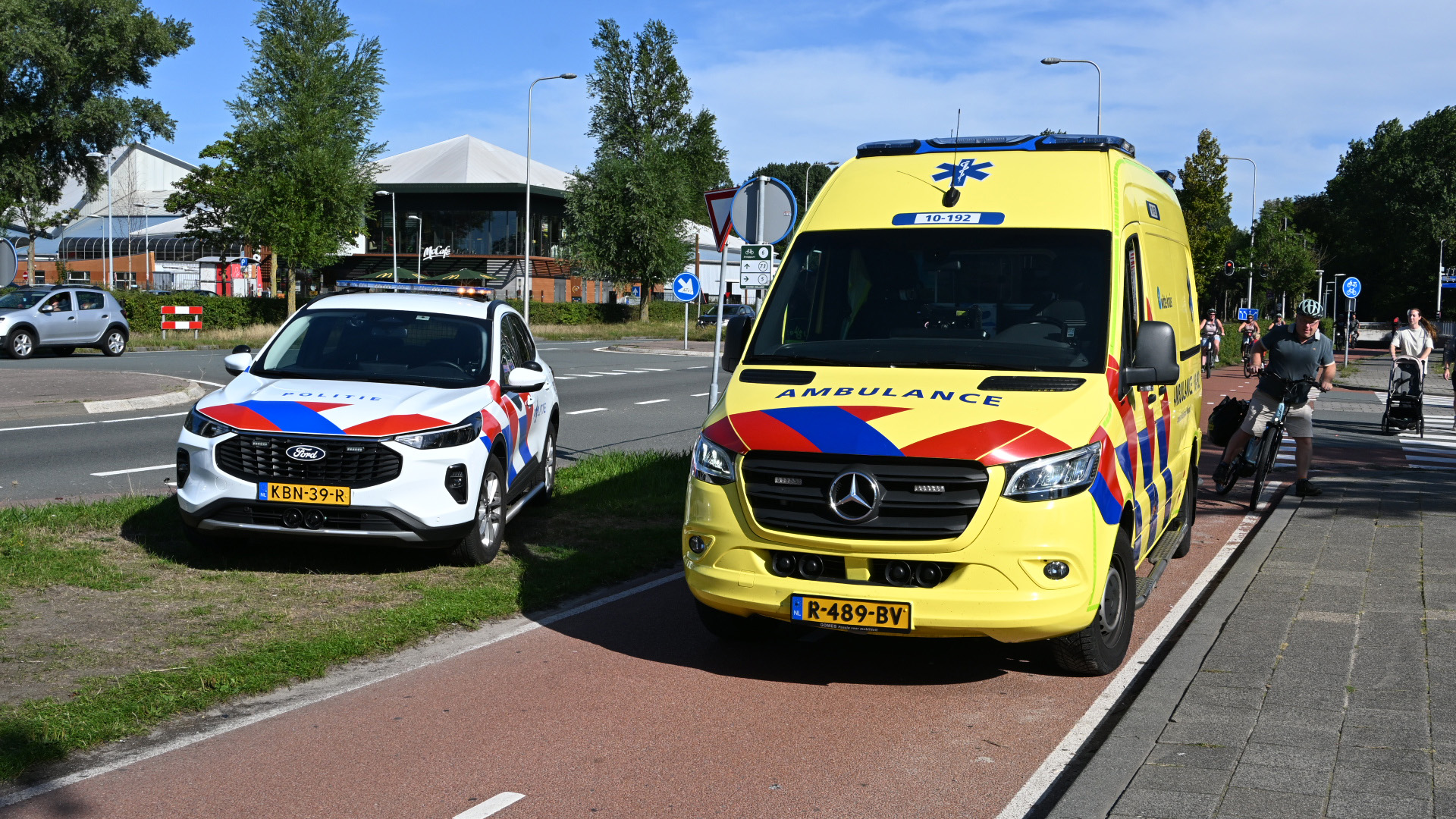 Gele ambulance en politieauto geparkeerd langs een fietspad; fietsers passeren op een zonnige dag