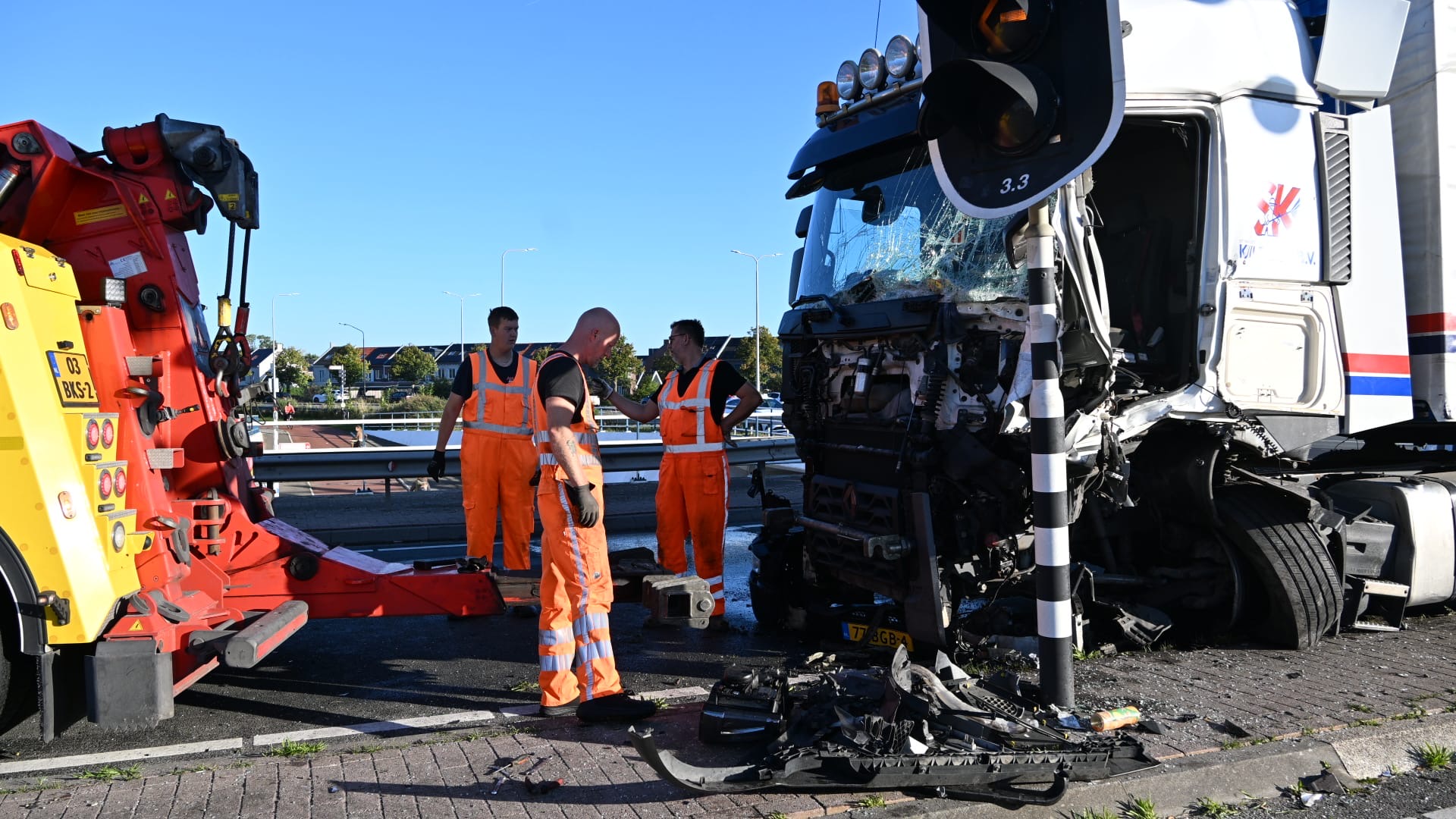 Wegwerkers in oranje pakken bij een zwaar beschadigde vrachtwagen na een ongeval, met takelwagen en brokstukken