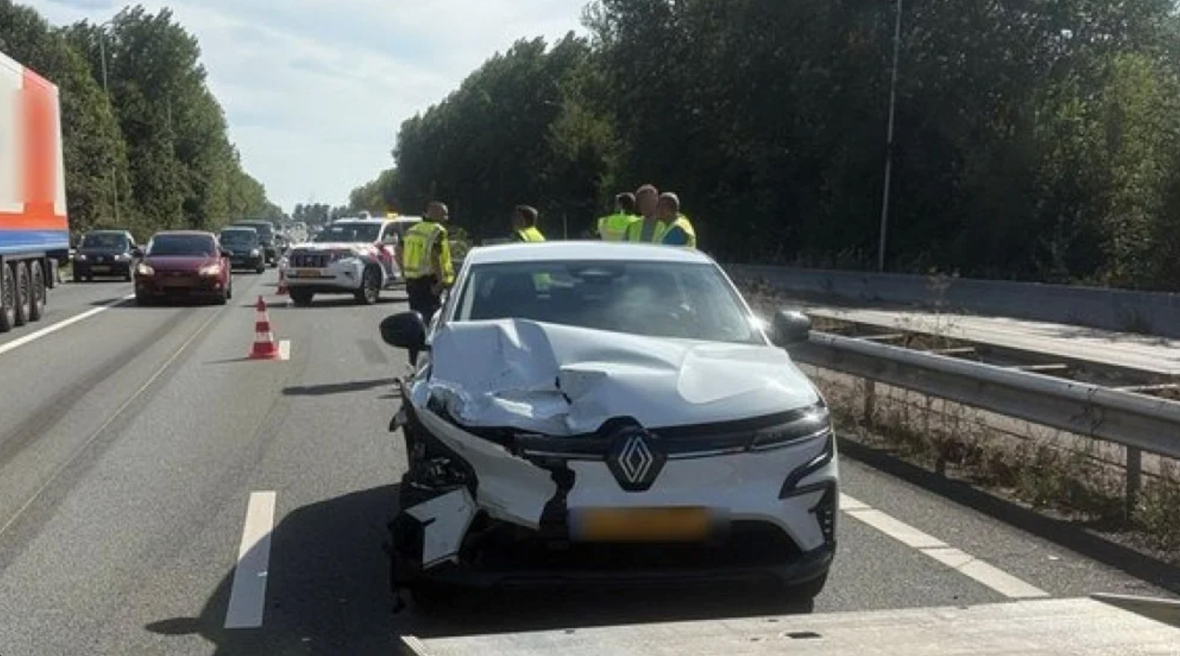Beschadigde witte auto na aanrijding op snelweg, met verkeerskegels en hulpdiensten in gele hesjes op de achtergrond