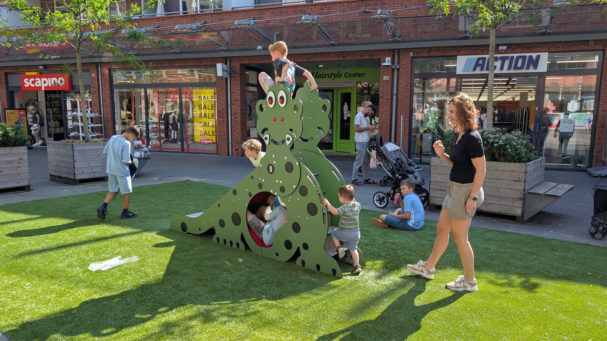 Kinderen spelen en klimmen op een groen speeltoestel in de vorm van een kikker op een plein met winkels