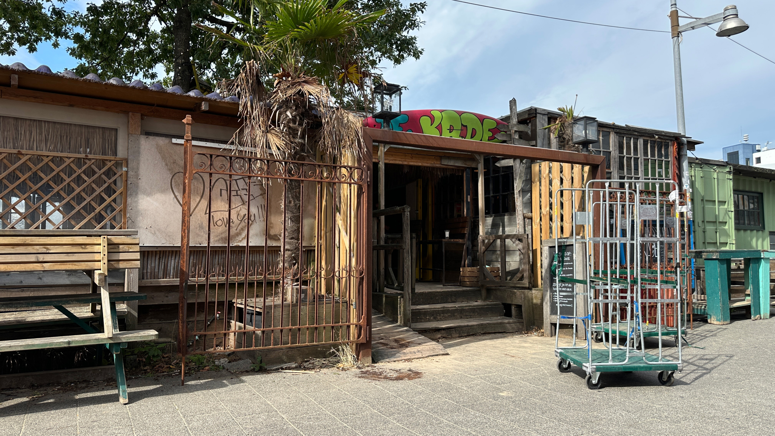 Rommelige buitenkant van houten bar met hek, palmboom, picknicktafel en rolkooi op het trottoir