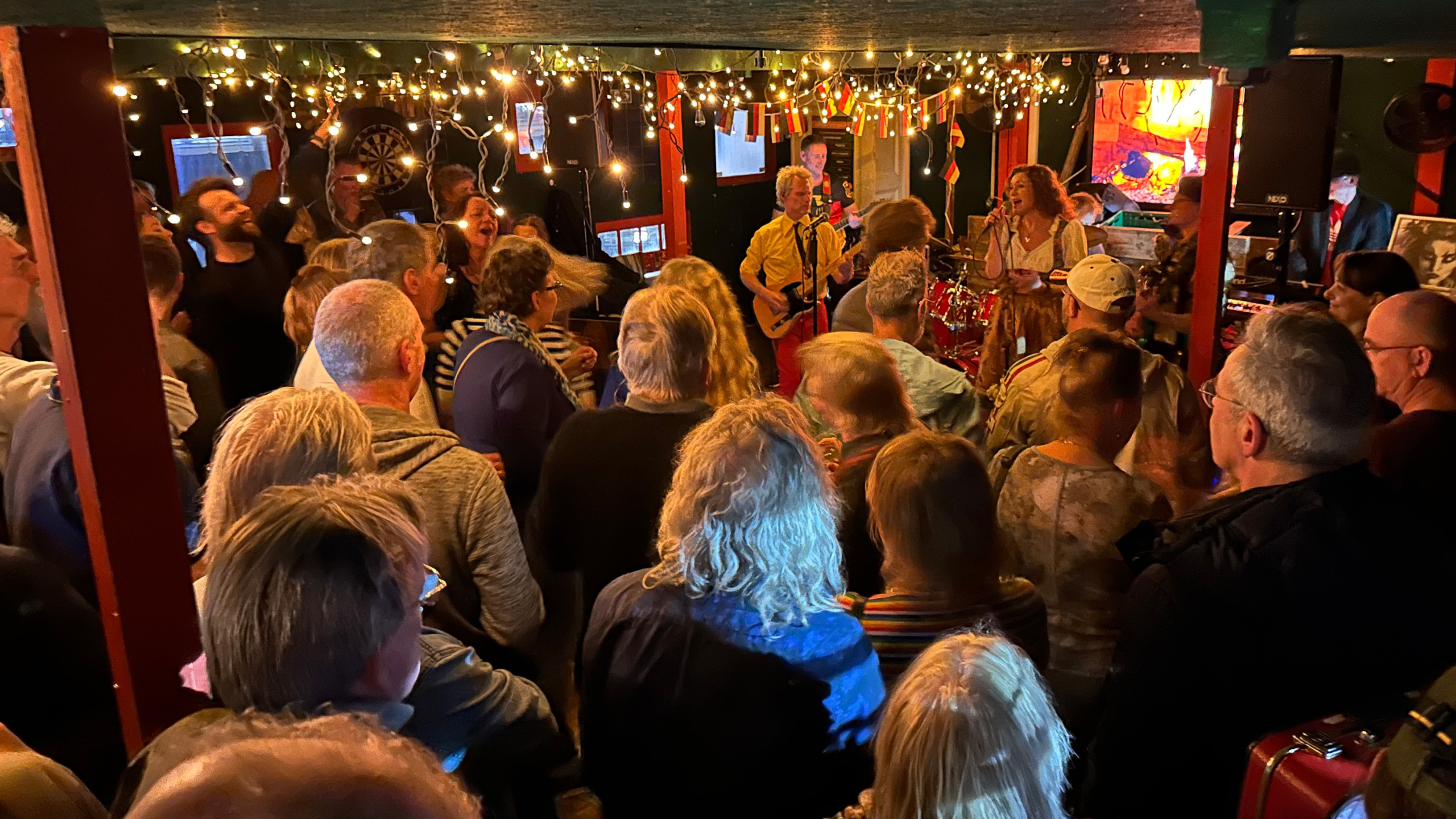 Drukke kroeg met liveband en zangeres; publiek staat dicht op elkaar onder sfeervolle lichtslingers en vlaggetjes