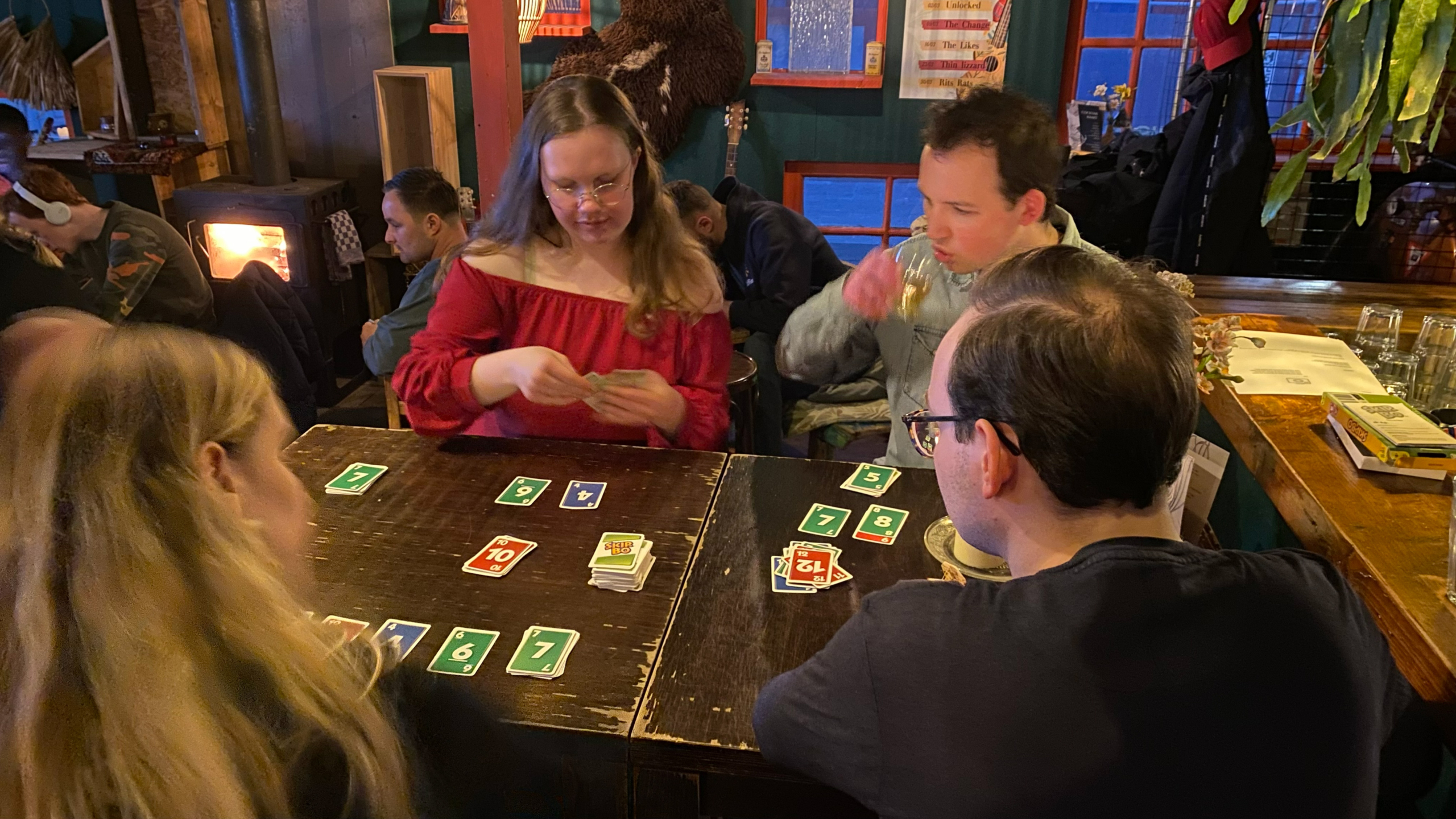 Groep vrienden speelt Skip-Bo aan een tafel in een gezellige kroeg, met kaarten op tafel en een houtkachel op de achtergrond