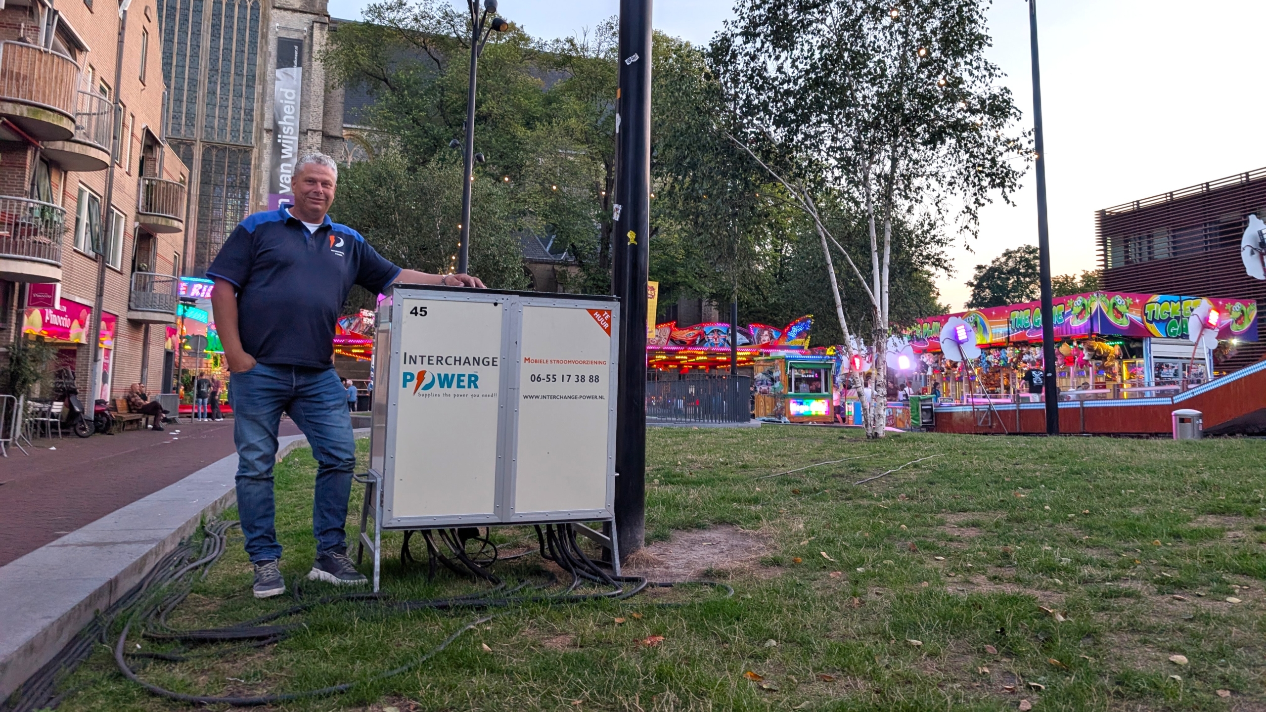 Man staat naast stroomkast met tekst Interchange Power, met kermisattracties en lichtjes op de achtergrond