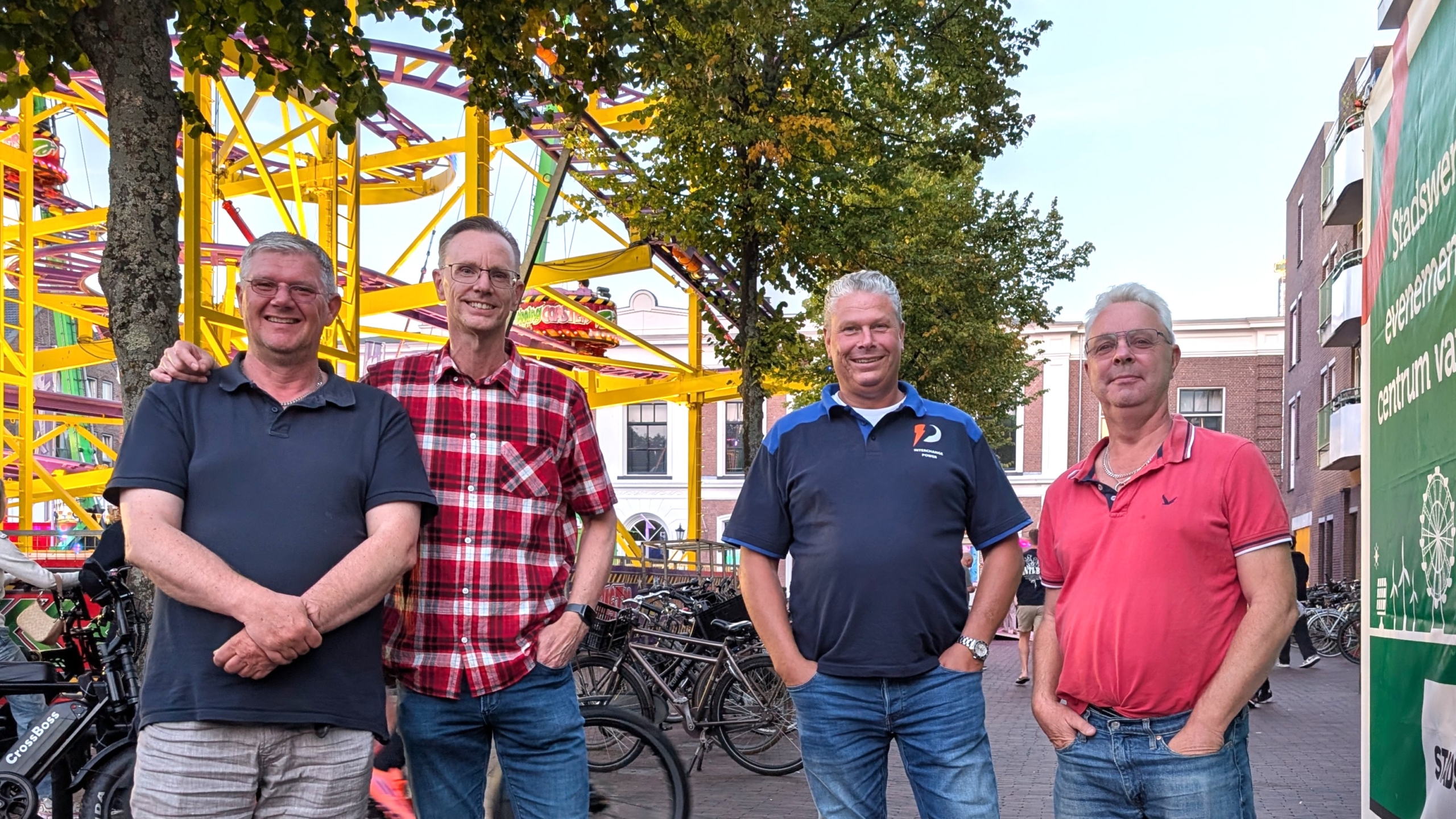 Vier mannen poseren voor een attractie op een plein, met fietsen op de achtergrond en bomen erboven