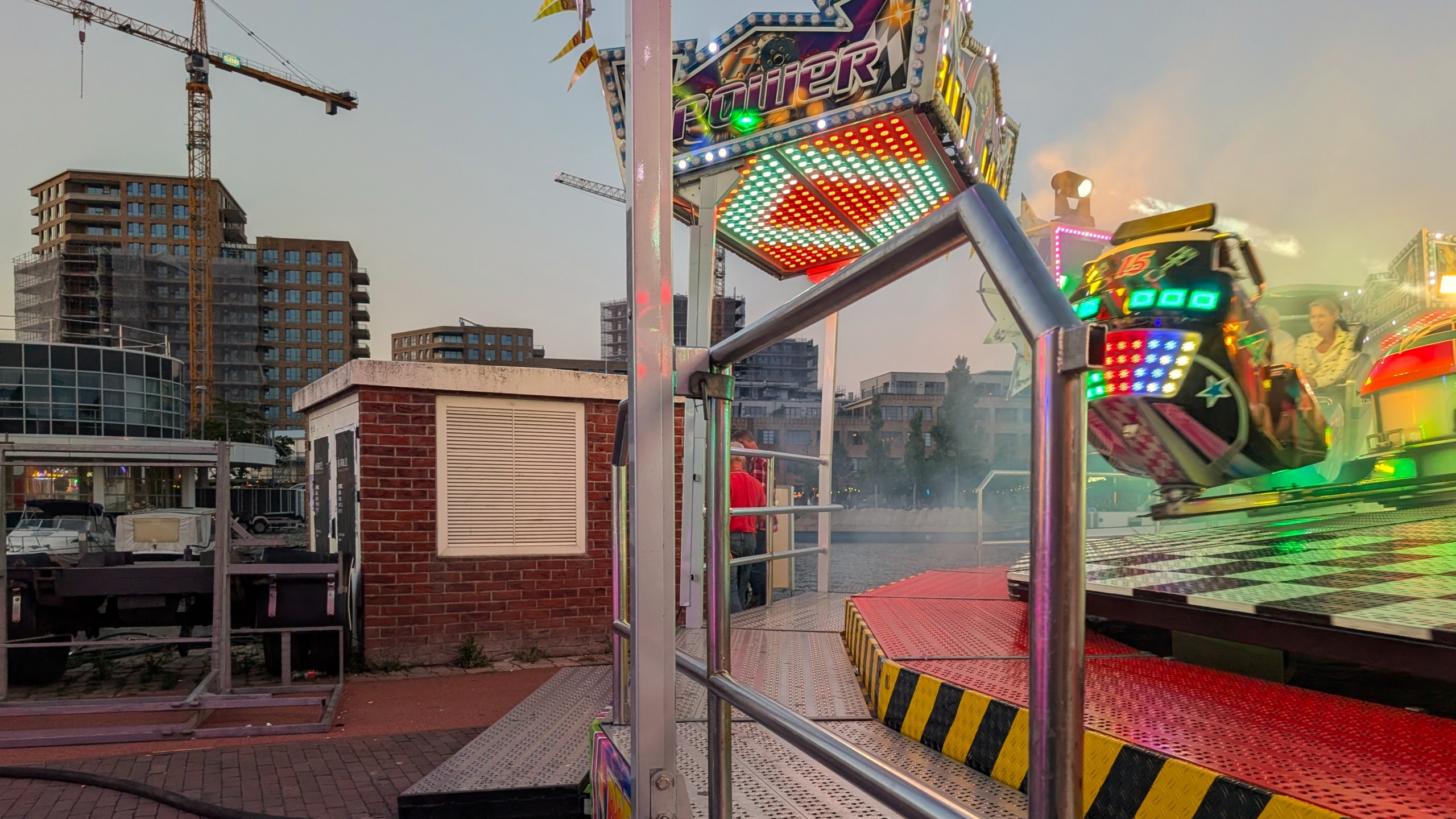 Kermisattractie met felle LED-lichten bij schemering, met bouwkranen en flats op de achtergrond