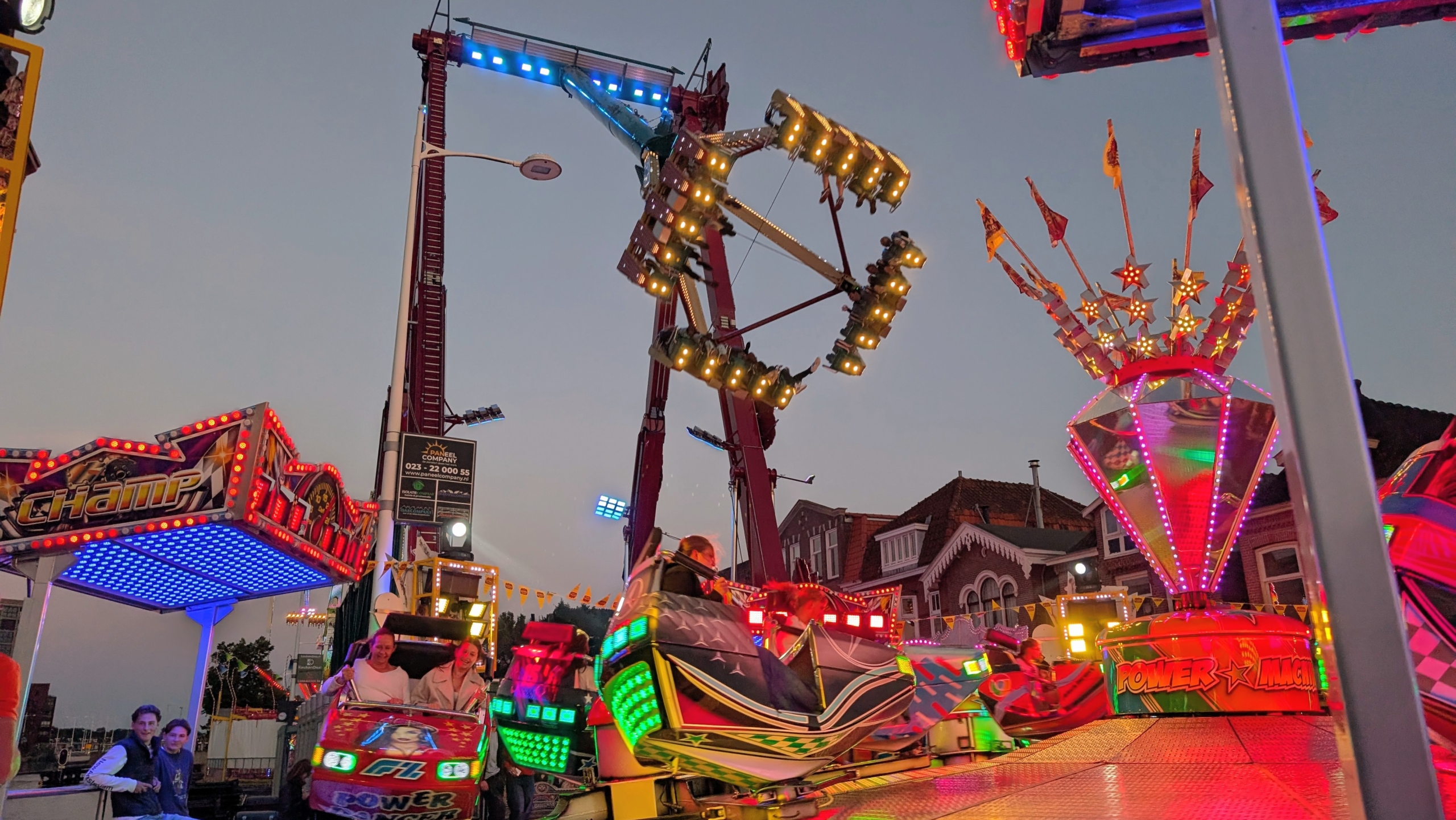 Verlichte kermisattracties bij schemering, met ronddraaiende thrillride op de achtergrond en botsauto’s op de voorgrond