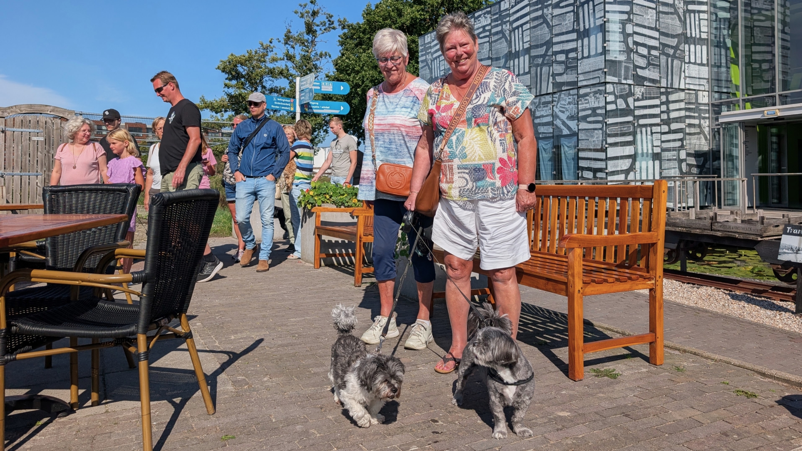 Twee vrouwen lopen met twee kleine honden aan de lijn over een terras, met mensen en een modern gebouw op de achtergrond