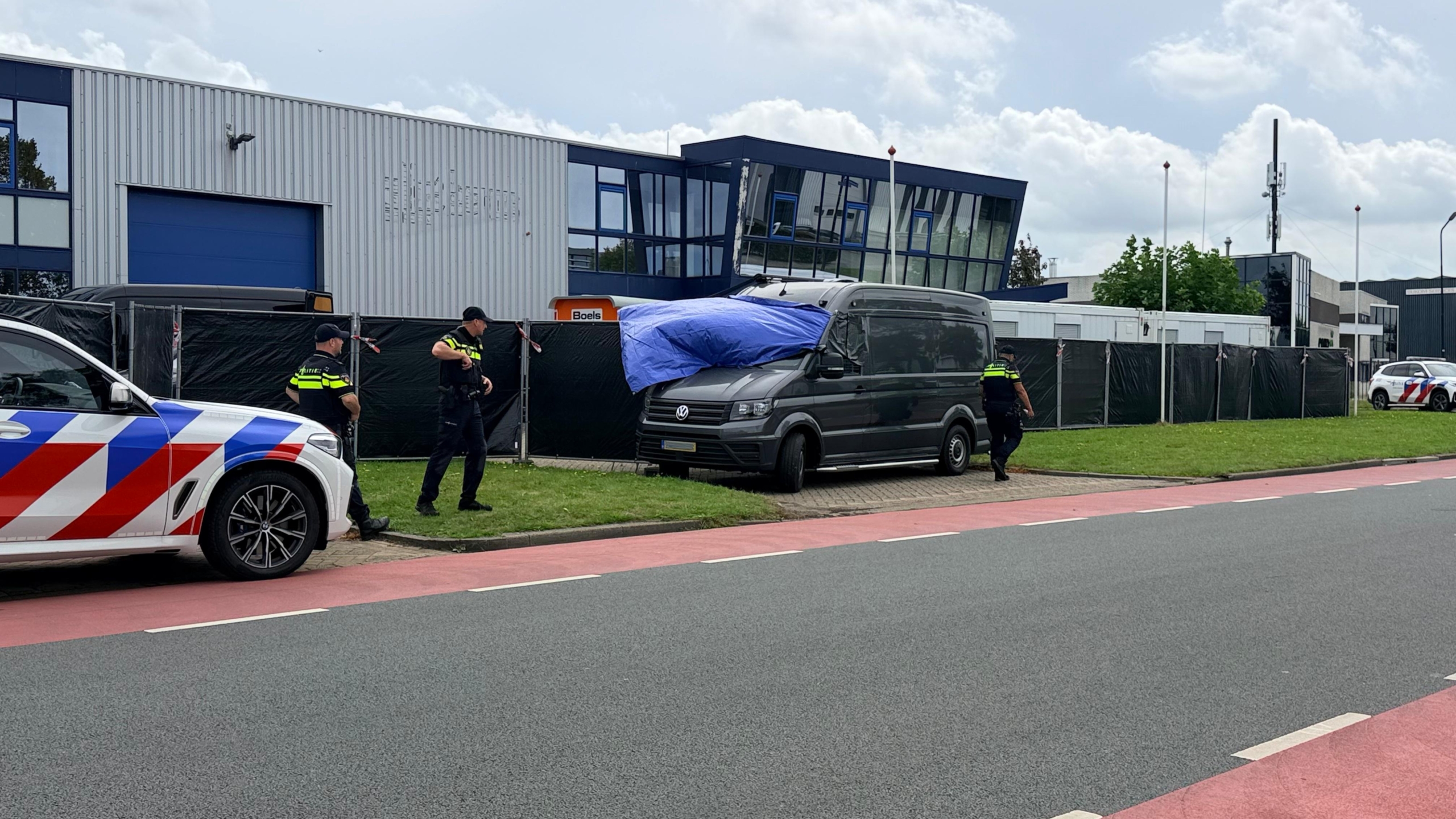 Politieagenten bij een grijze bestelbus met blauw zeil op een bedrijventerrein, met politieauto op de voorgrond
