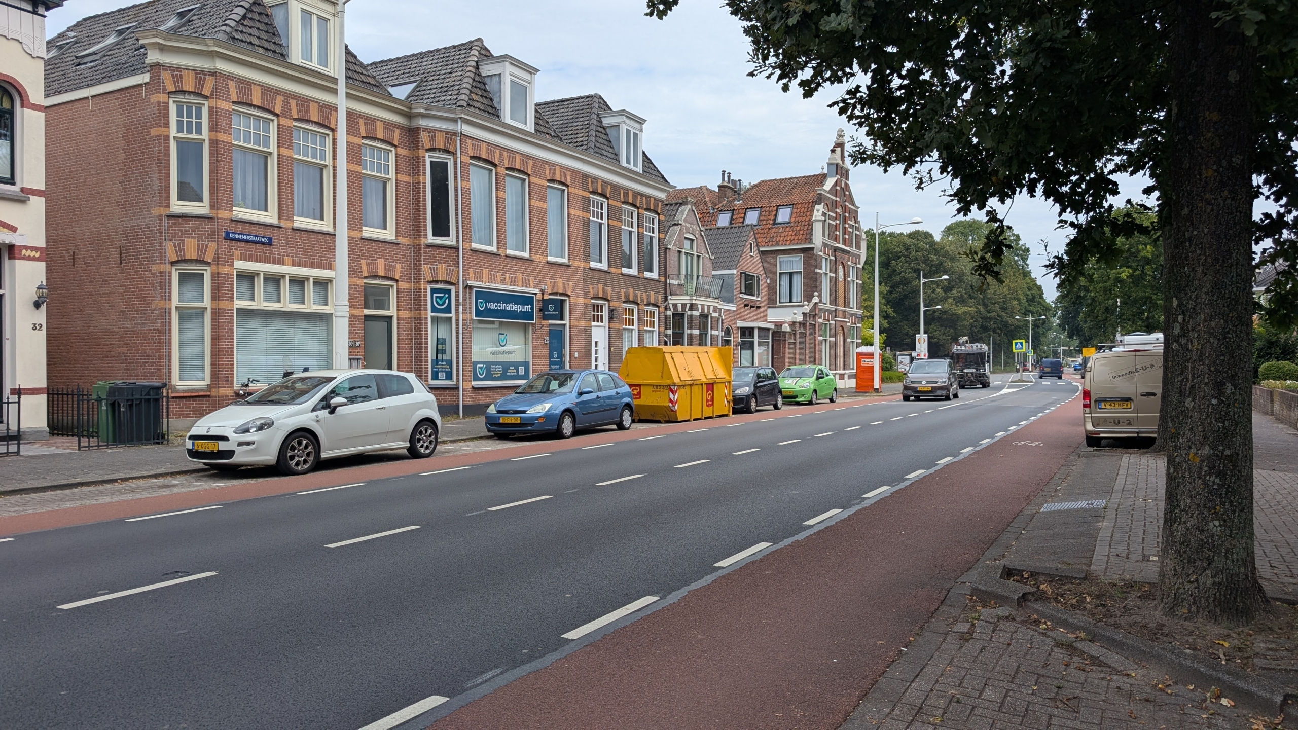 Straat met bakstenen panden, geparkeerde auto’s en gele container; gevelbord met tekst “vaccinatiepunt”