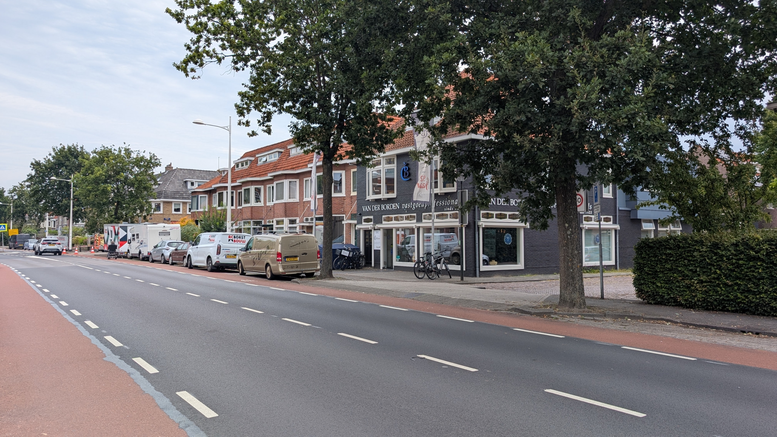 Straat met fietsstrook en geparkeerde busjes, langs bomen en winkelpanden met bord “Van der Borden”