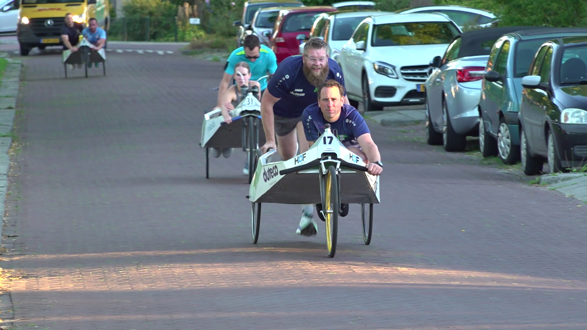 Twee mannen duwen een driewielige ligfiets met startnummer 17 door een straat met geparkeerde auto’s