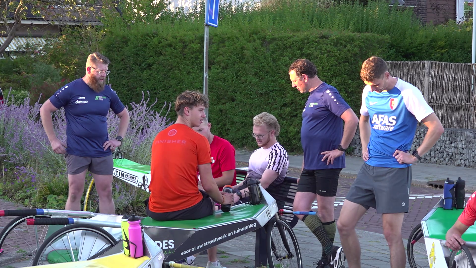 Groep mannen in sportkleding bij roeifietsen, in gesprek langs de straat met groen op de achtergrond