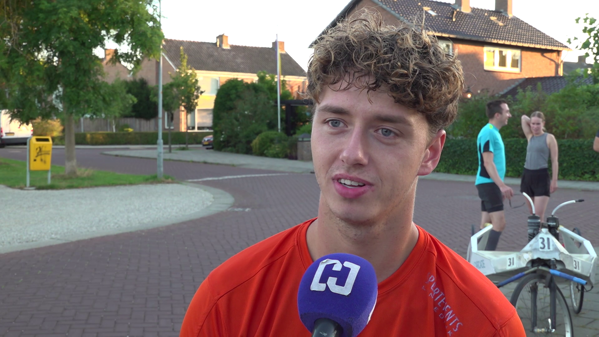 Jonge man met krullend haar wordt buiten in woonwijk geïnterviewd met paarse microfoon; sporters en fiets op achtergrond