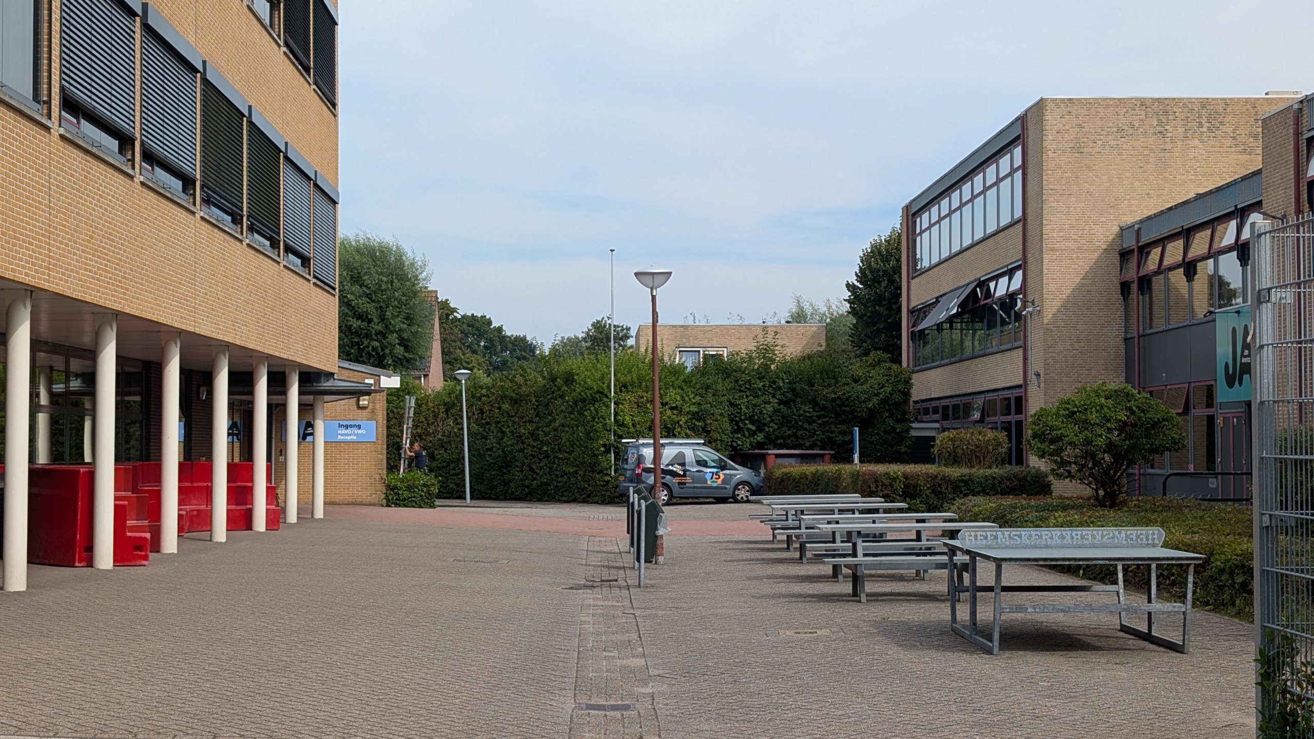 Schoolplein tussen twee bakstenen gebouwen met picknicktafels, lantaarnpaal en geparkeerde bestelbus bij groen struikgewas