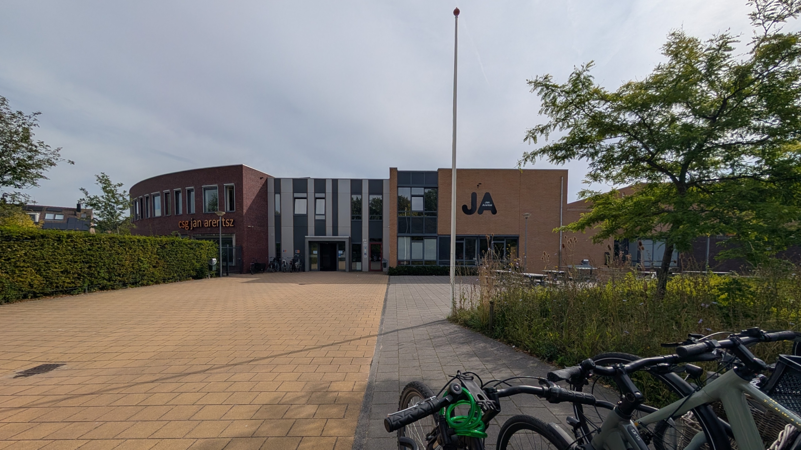 Schoolgebouw met opschrift CSG Jan Arentsz, met vlaggenmast op het plein en fietsen op de voorgrond