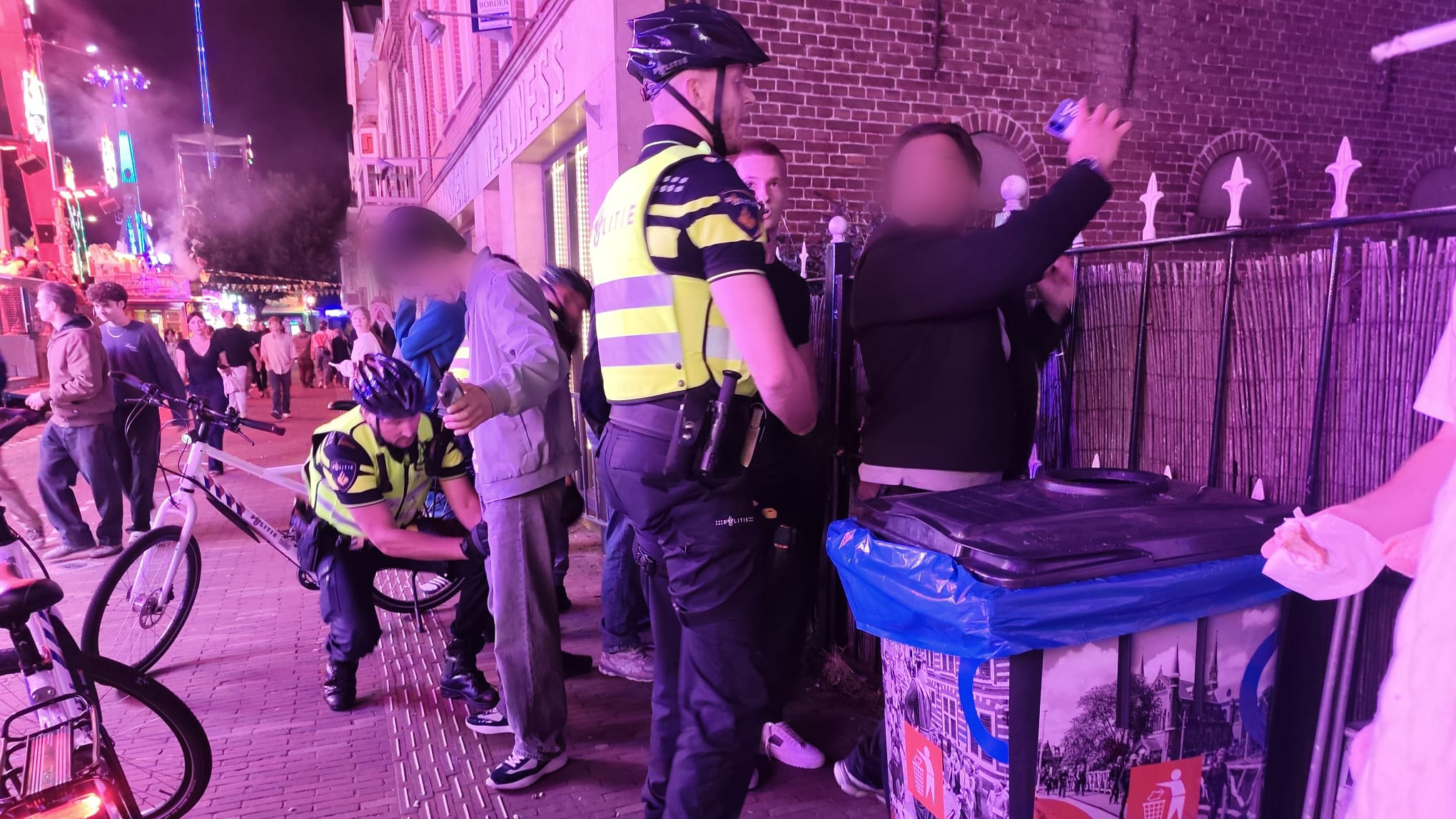 Politie fouilleert twee mannen op een drukke, felverlichte straat in de avond, met fietsen en omstanders op de achtergrond
