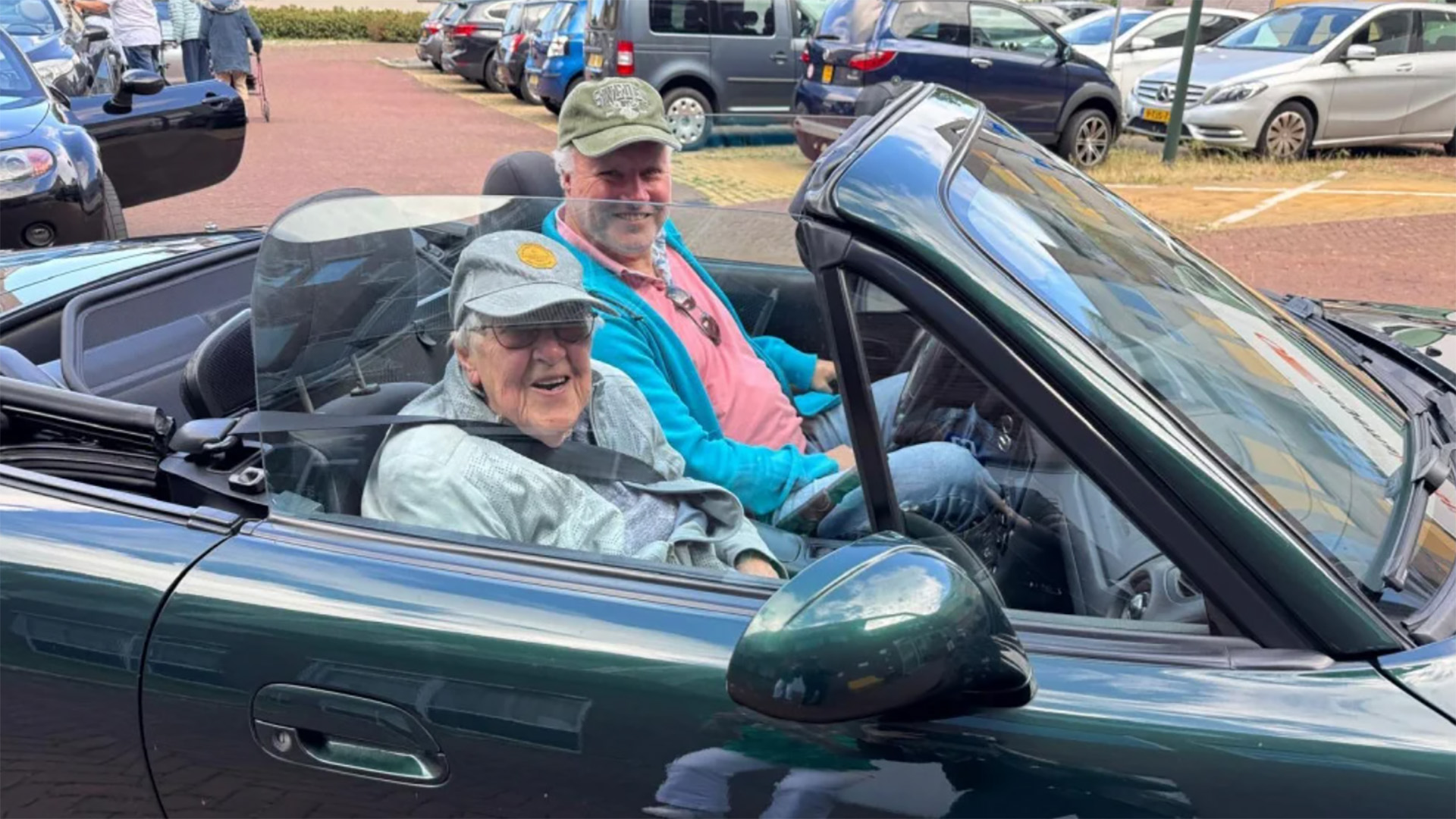Twee mannen glimlachen in een donker groene cabriolet met open dak, geparkeerd tussen andere auto’s