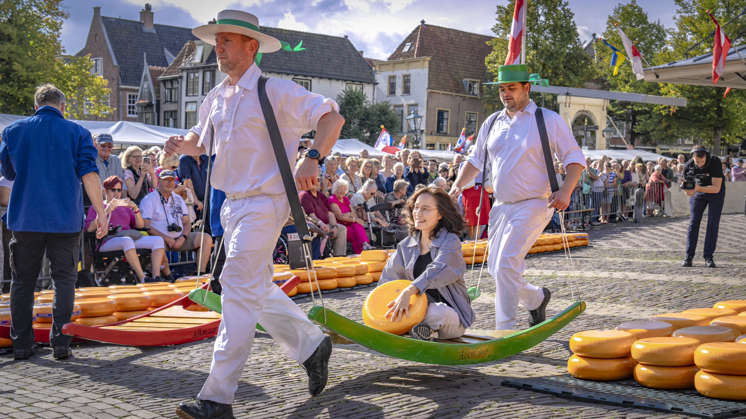 Kaasdragers in wit dragen een brancard met vrouw en kaaswiel, met publiek op een plein vol kaaswielen