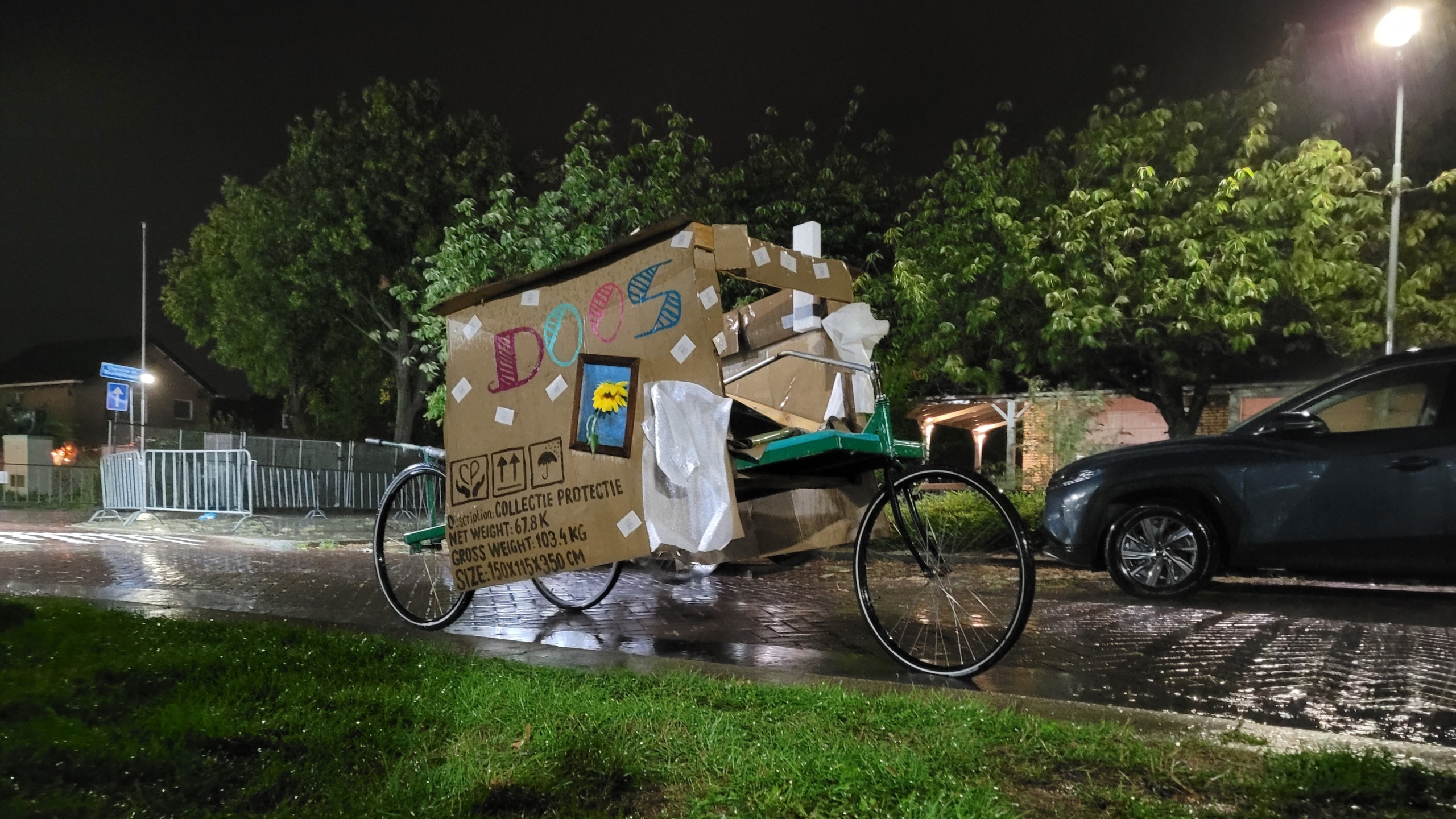 Bakfiets met kartonnen huisje erop, met de tekst “DOOS”, staat ’s nachts in de regen langs de weg