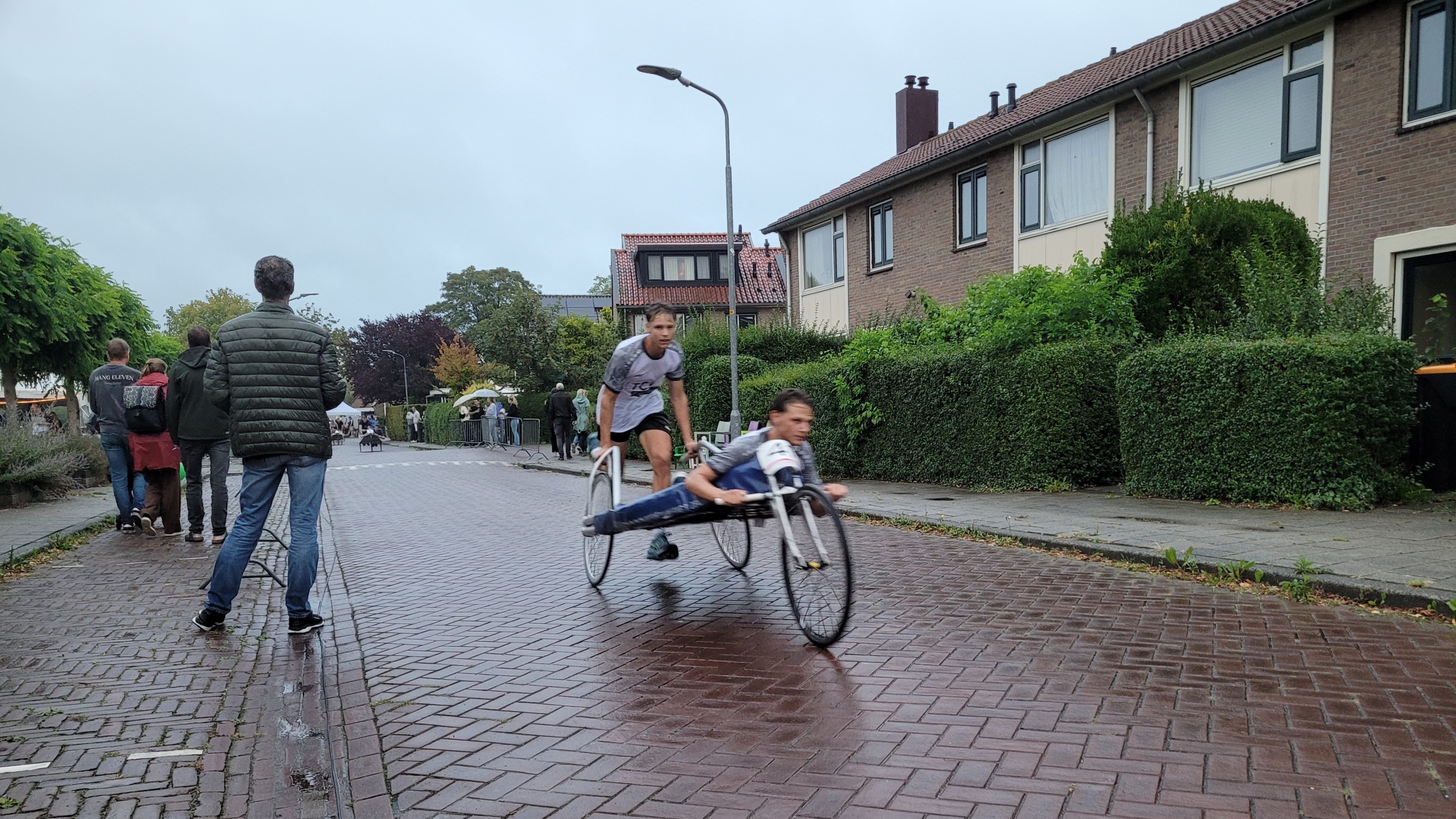 Handbiker racet over natte straat, gevolgd door hardloper; toeschouwers staan langs de weg bij woonhuizen