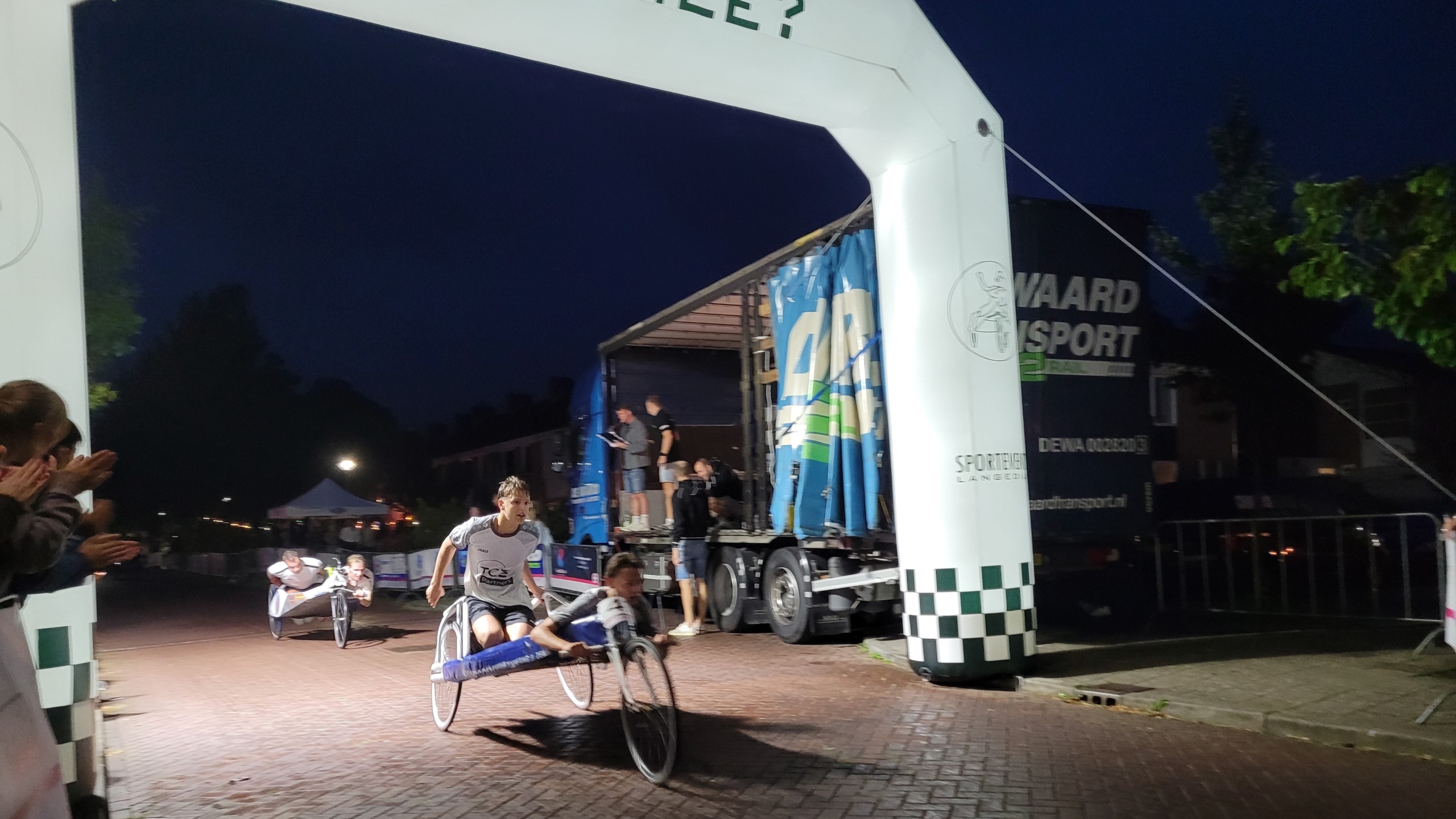 Twee handbikers finishen ’s avonds onder een opblaasbare finishboog, terwijl toeschouwers langs de weg applaudisseren