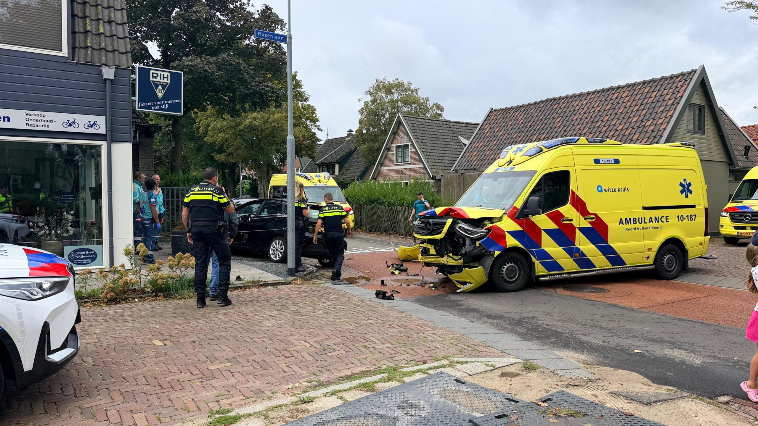 Beschadigde ambulance en politie bij een ongeluk op een kruispunt, met een zwarte auto en brokstukken op de weg