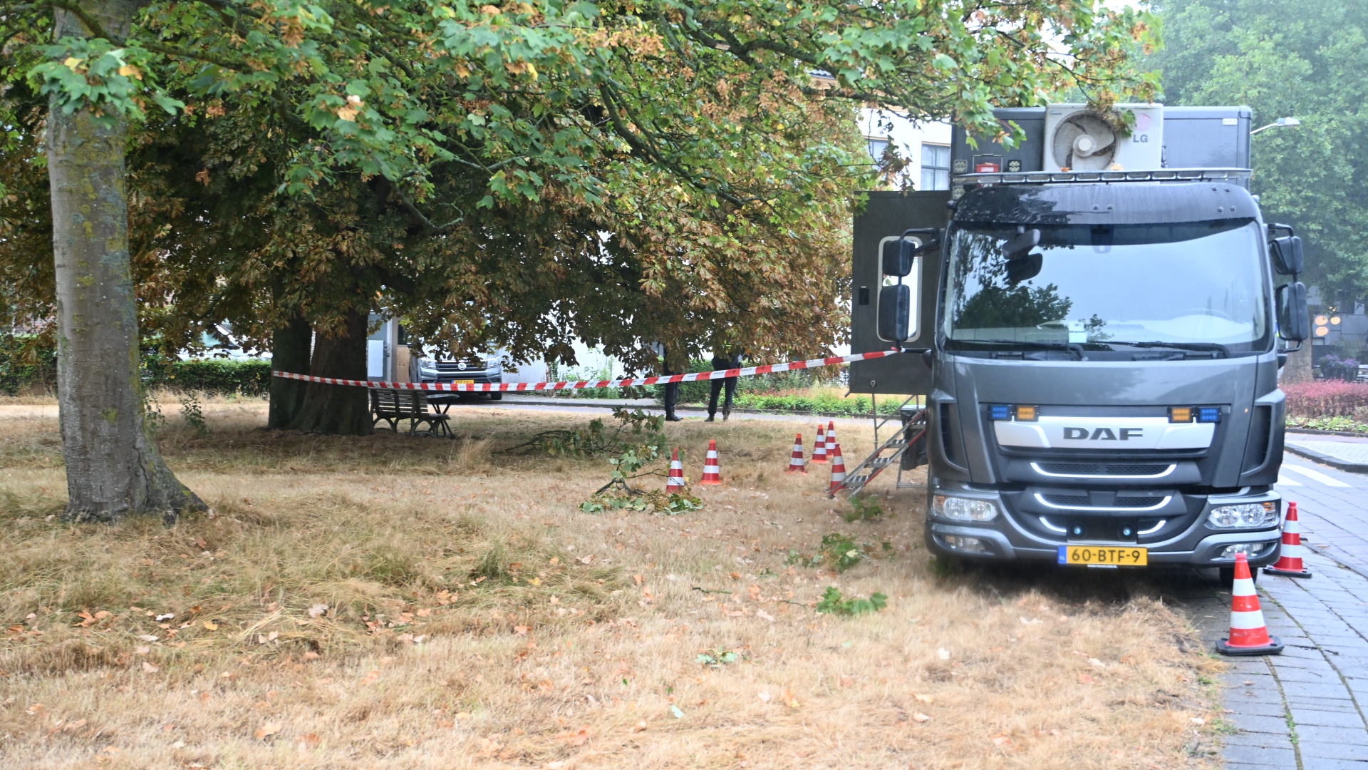 DAF-vrachtwagen bij afgezet gebied met verkeerskegels en rood-wit lint onder bomen naast een parkpad