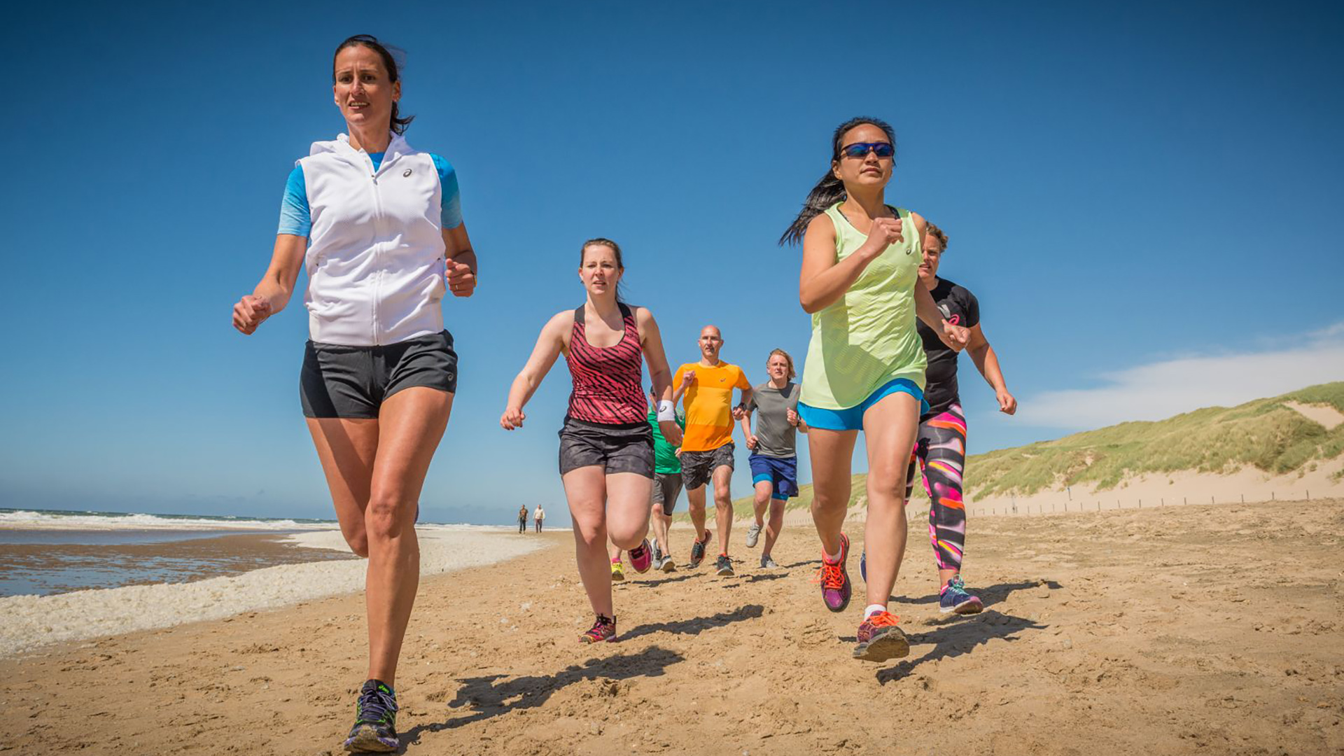 Groep hardlopers rent over het strand langs de zee en duinen onder een heldere blauwe lucht