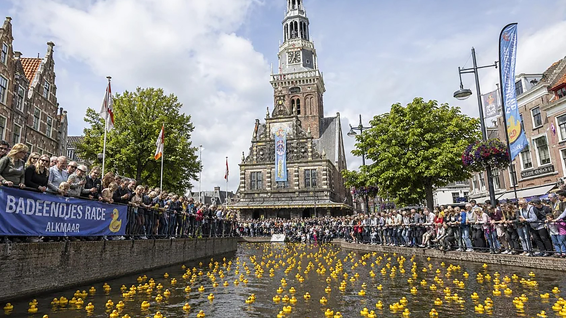 Toeschouwers langs een gracht bij het Waaggebouw in Alkmaar, met honderden gele badeendjes voor de Badeendjesrace