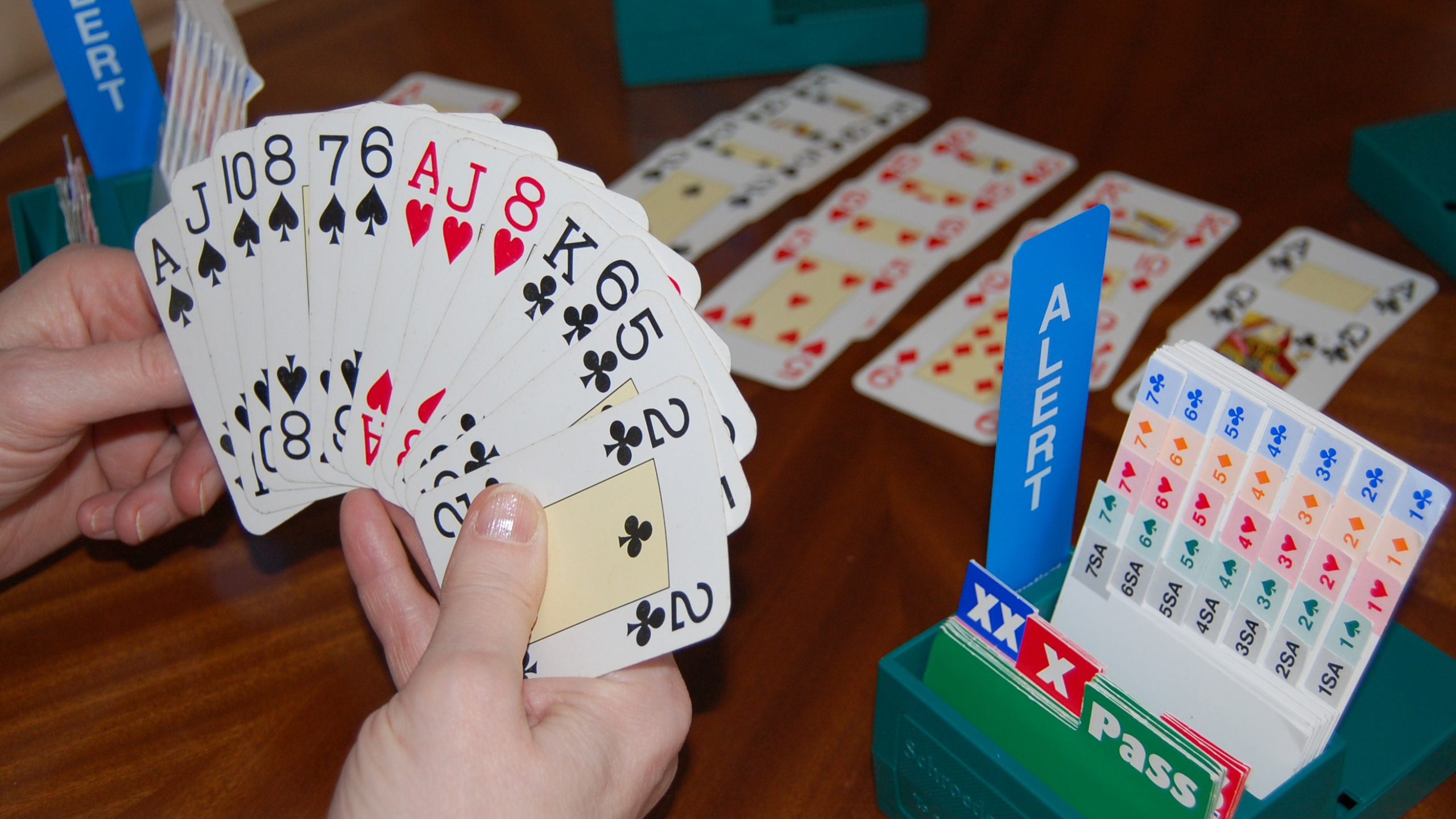 Hand met speelkaarten en bridge-biedbox op tafel, met uitgelegde kaarten op de achtergrond