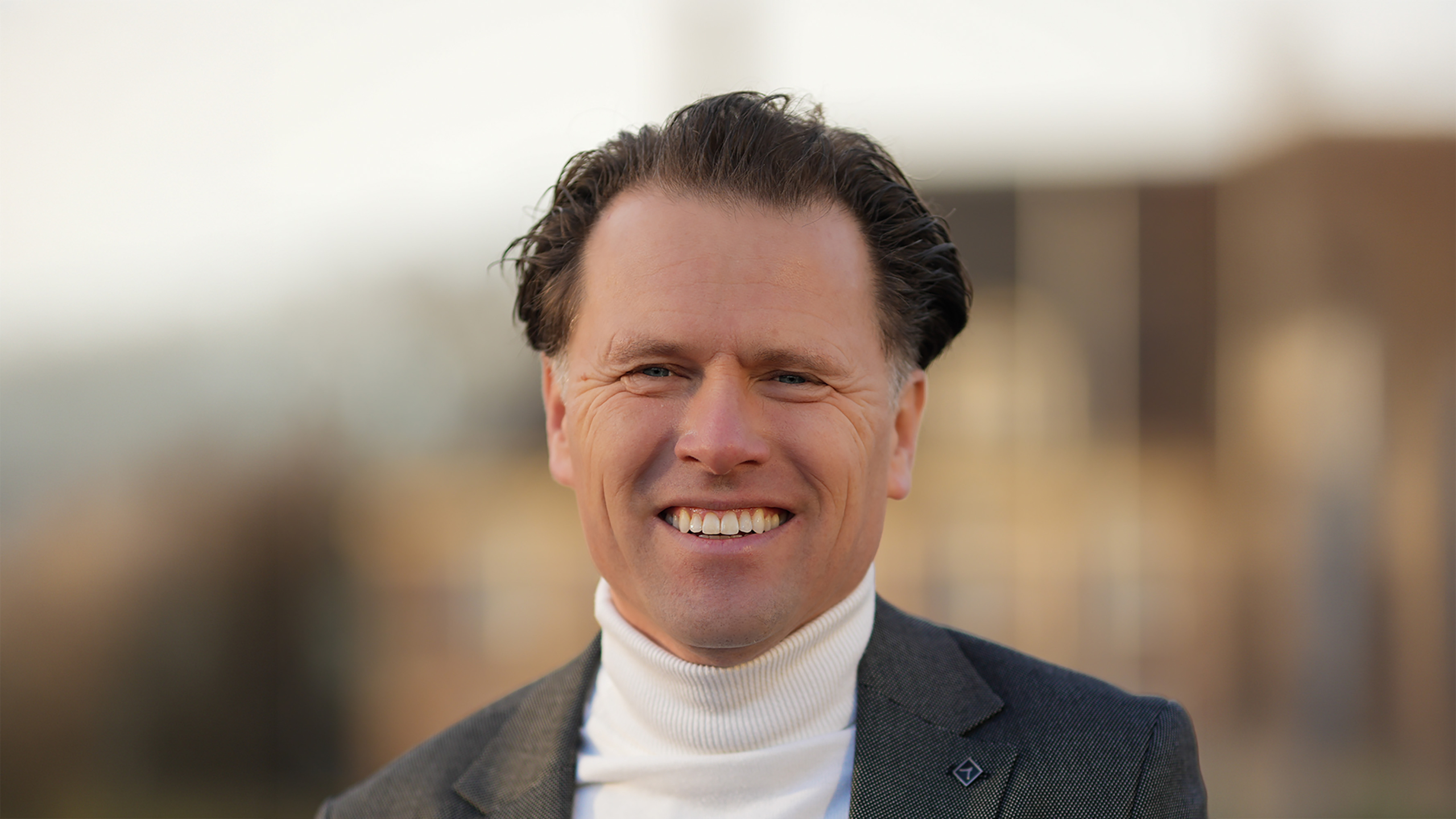 Portret van een glimlachende man in coltrui en colbert, met een wazige achtergrond van gebouwen