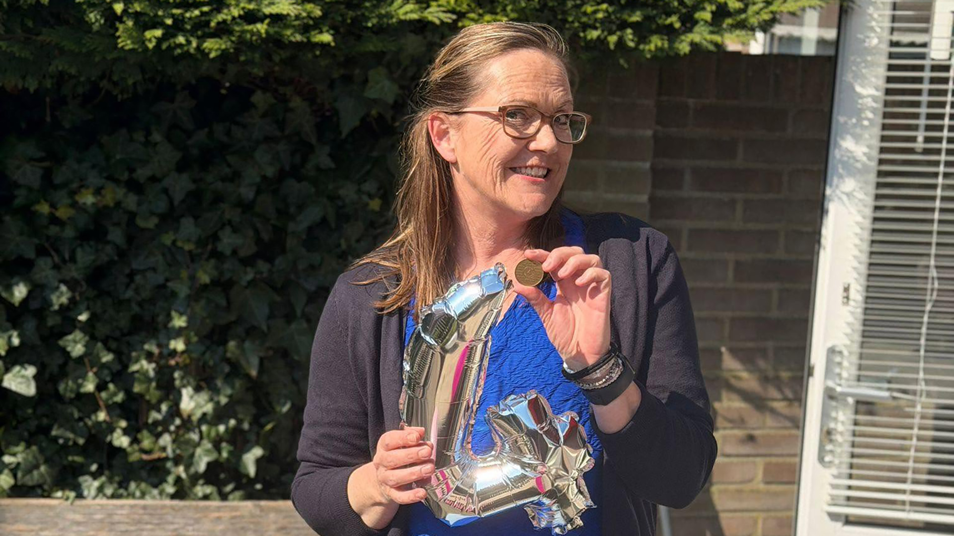 Vrouw met bril houdt een gouden medaille en een zilveren ballon in de vorm van 40 vast in een zonnige tuin