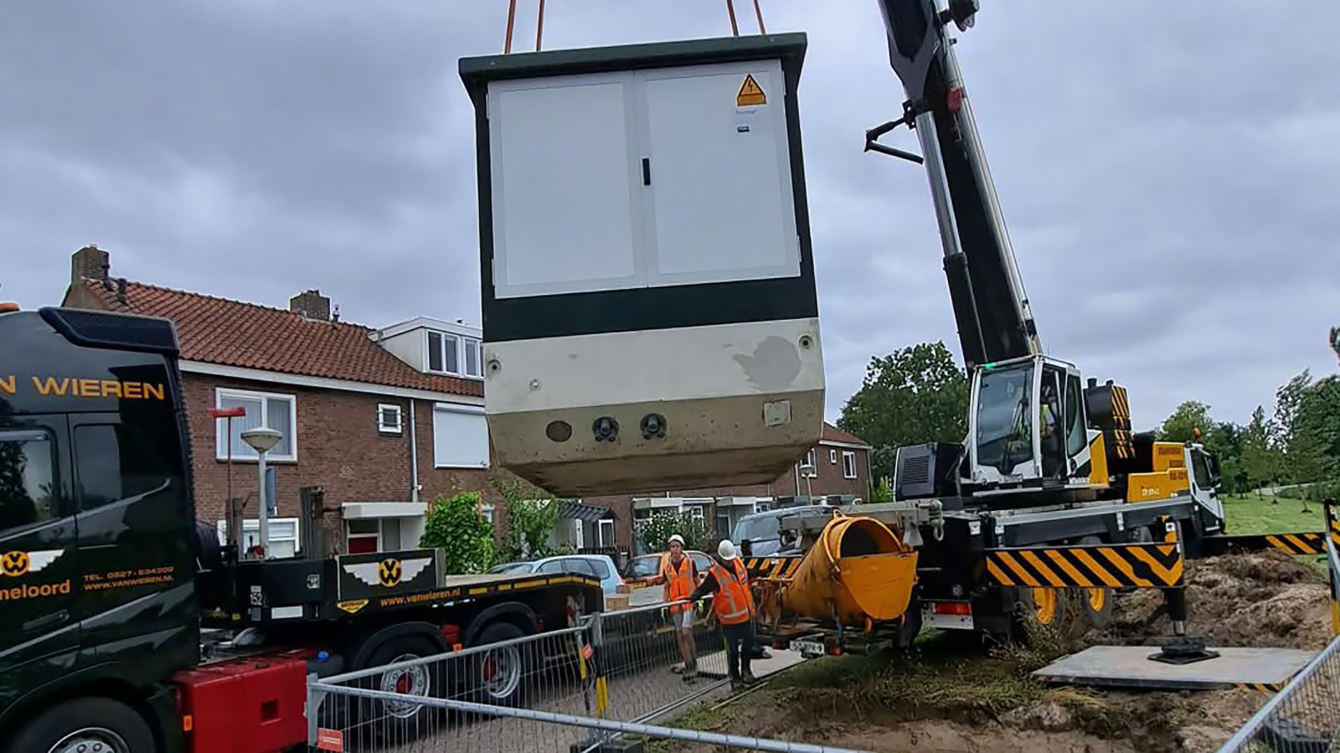 Kraanwagen hijst een grote prefab-unit boven een bouwplaats, met twee bouwvakkers in oranje hesjes erbij