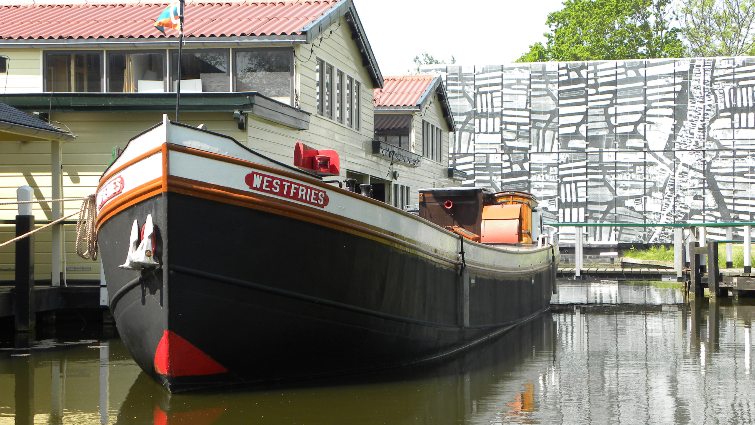 Binnenvaartschip Westfries aangemeerd bij houten gebouwen aan het water, met grote zwart-witte muurschildering op de achtergrond
