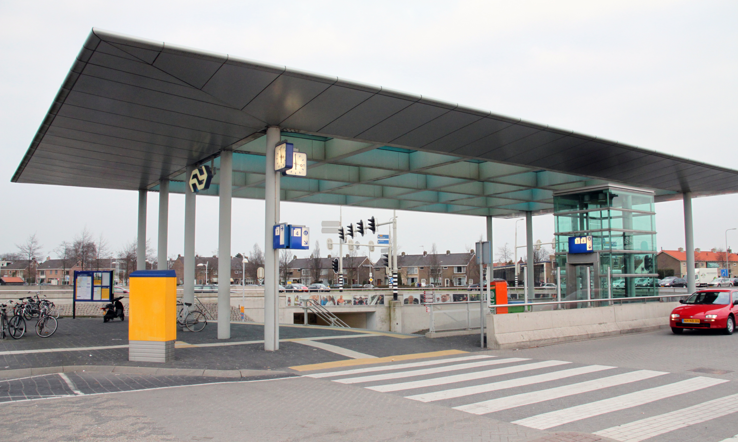 Overkapping van een stationstraverse met klok, borden en glazen lift, met fietsenrekken en een zebrapad op de voorgrond